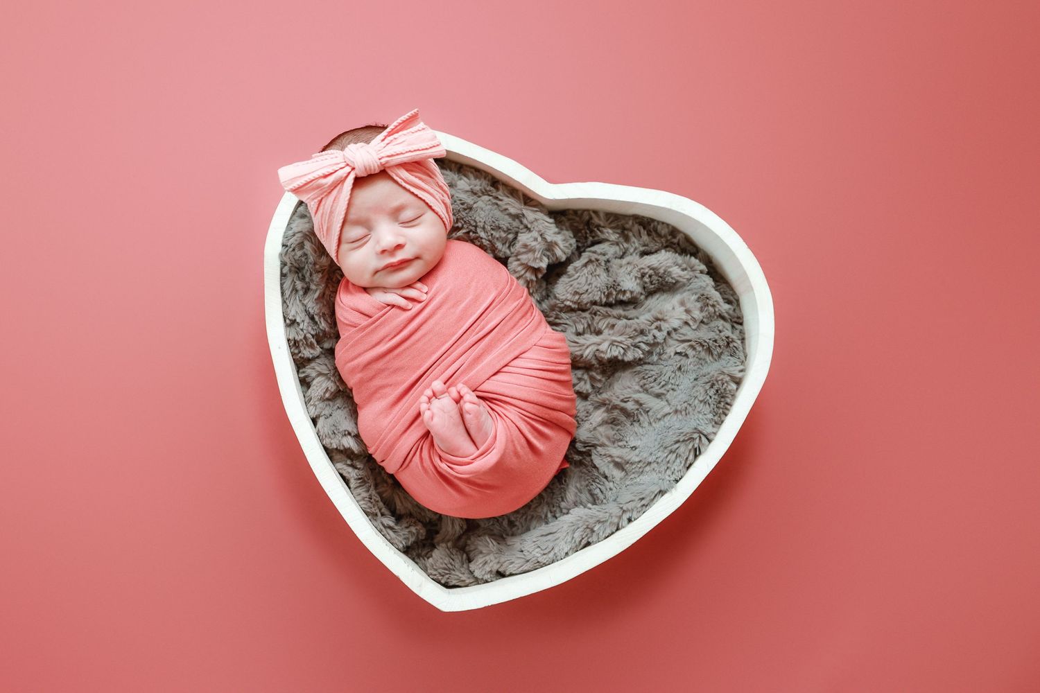 Peaceful newborn baby wrapped in coral pink swaddle with bow headband resting in heart-shaped prop.