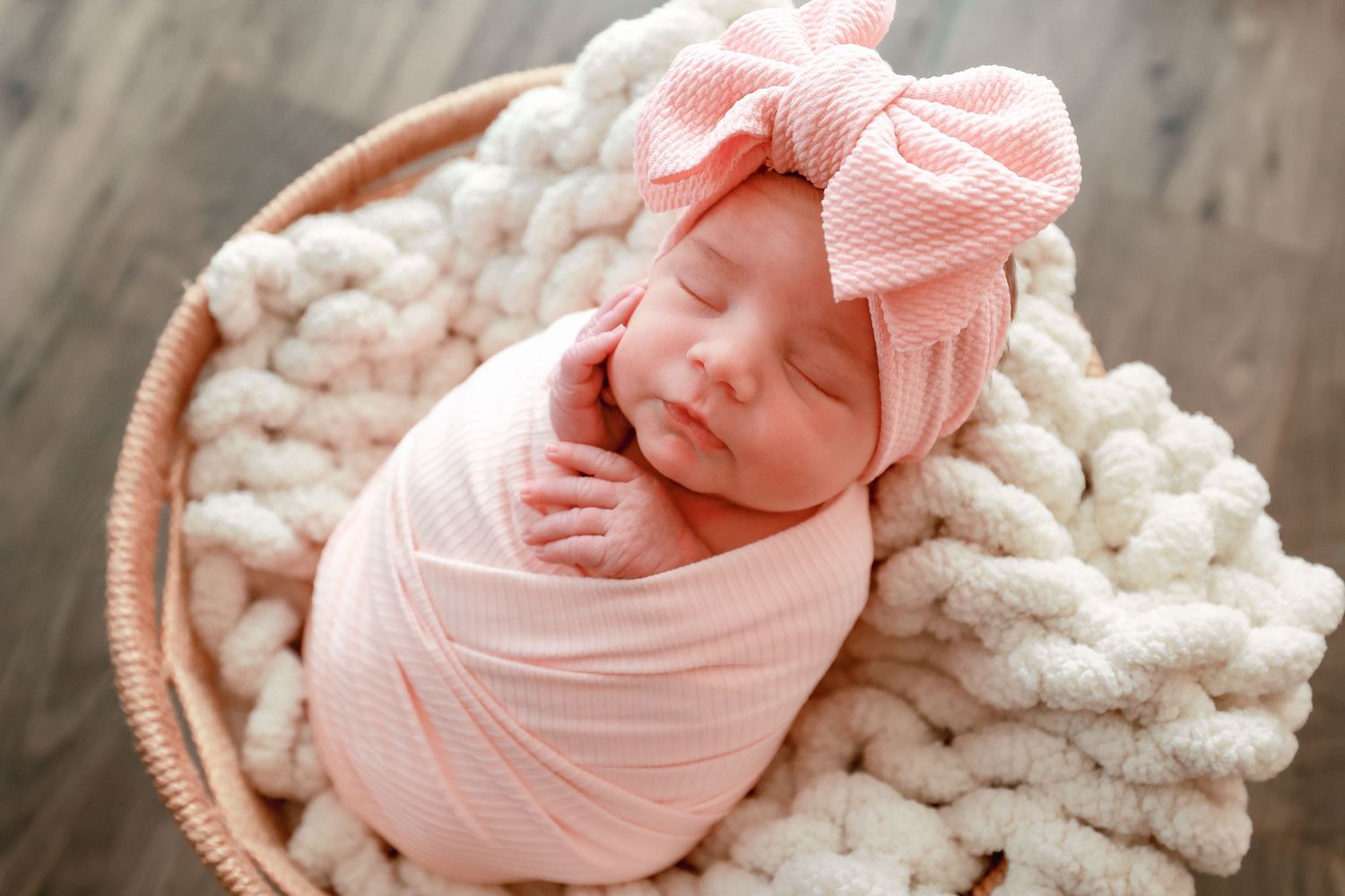 Newborn baby girl swaddled in pink wrap with bow headband sleeping peacefully in a wicker basket.