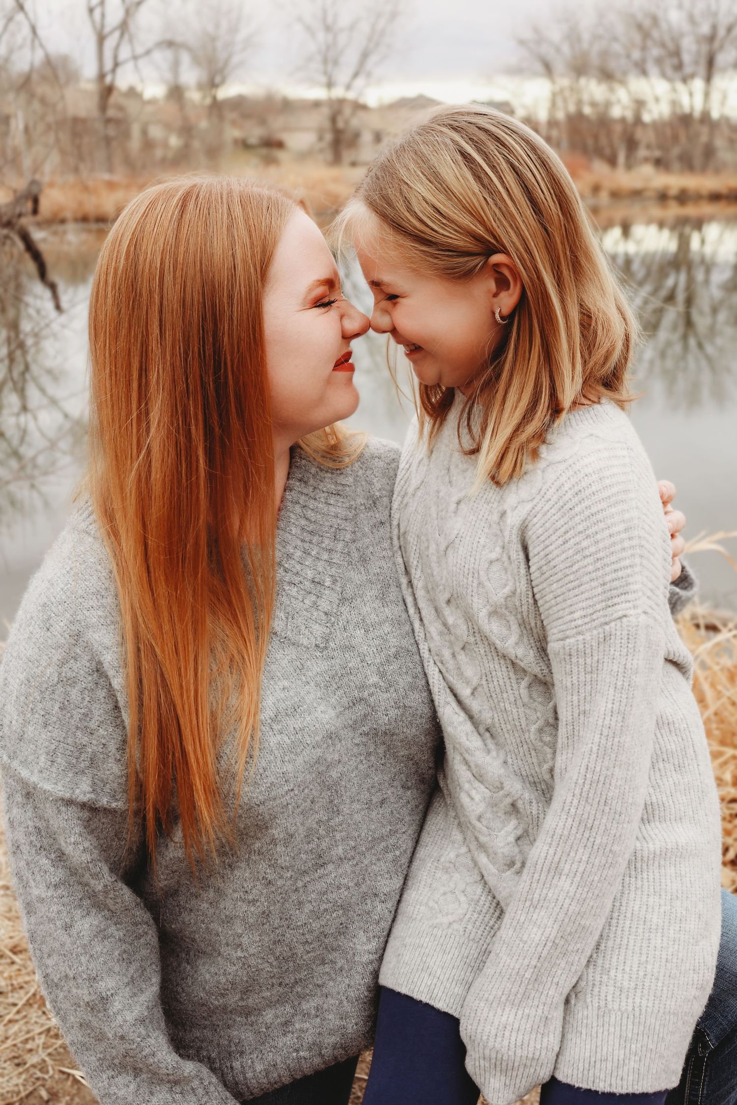 Two girls in matching gray sweaters touching noses and smiling by a reflective pond.