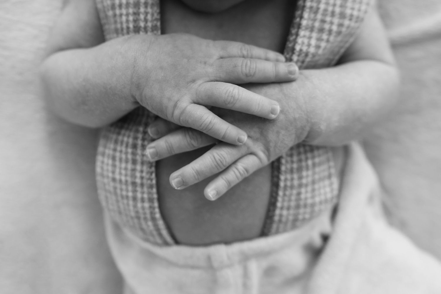 Black and white close-up of a newborn baby's tiny hands resting on chest in a plaid wrap.
