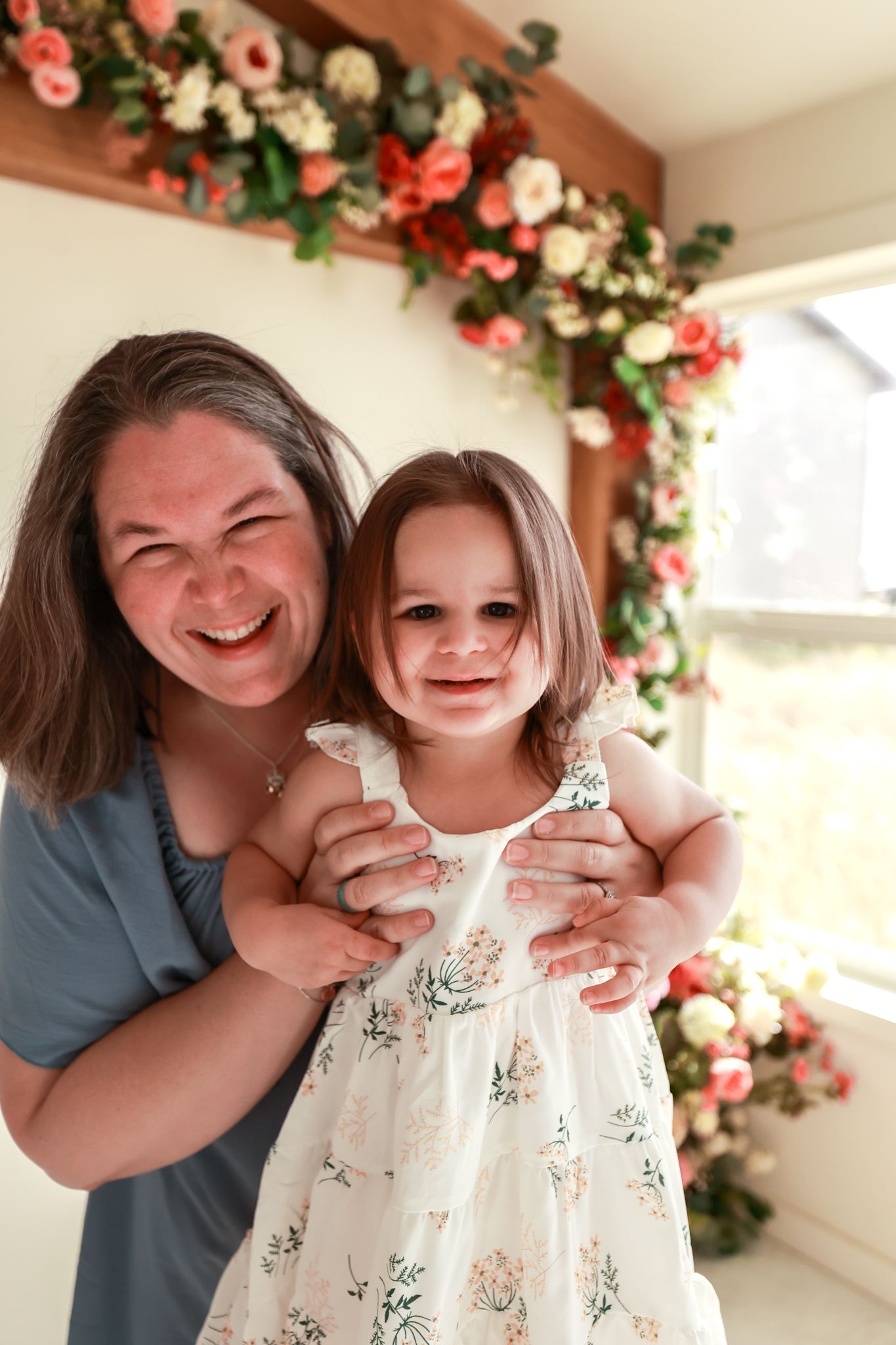 A laughing woman hugs a smiling little girl in a floral dress, posing before a beautiful flower garland backdrop.