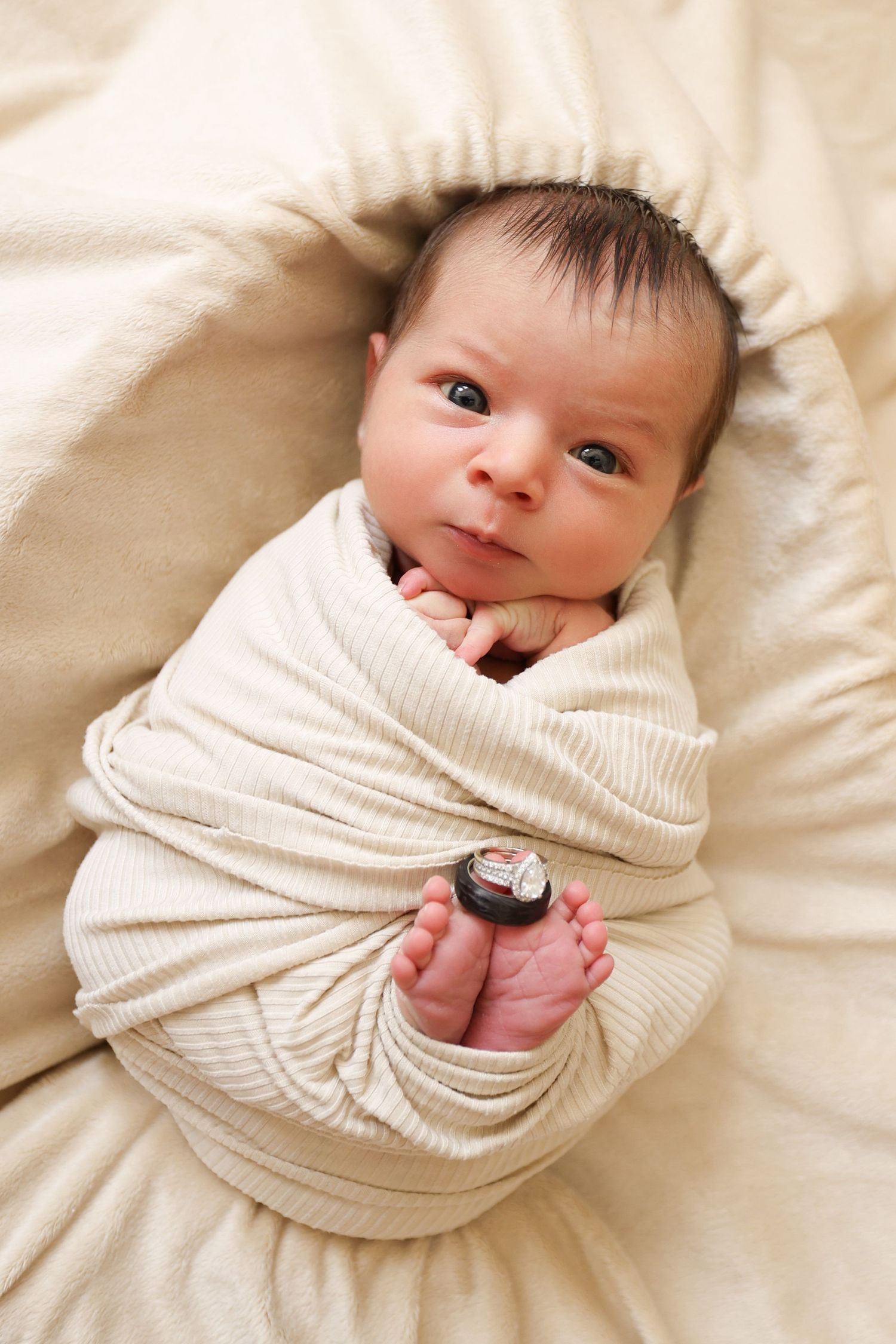 Wide-eyed newborn baby snugly swaddled in cream ribbed wrap with tiny feet peeking out in bassinet.