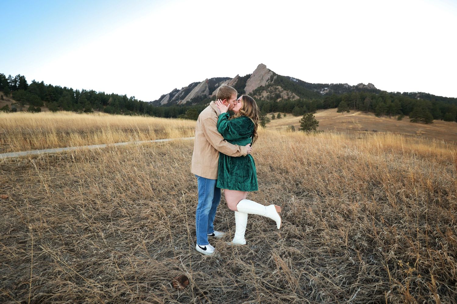Couple embracing and kissing in golden field with dramatic Flatiron rock formations behind them.
