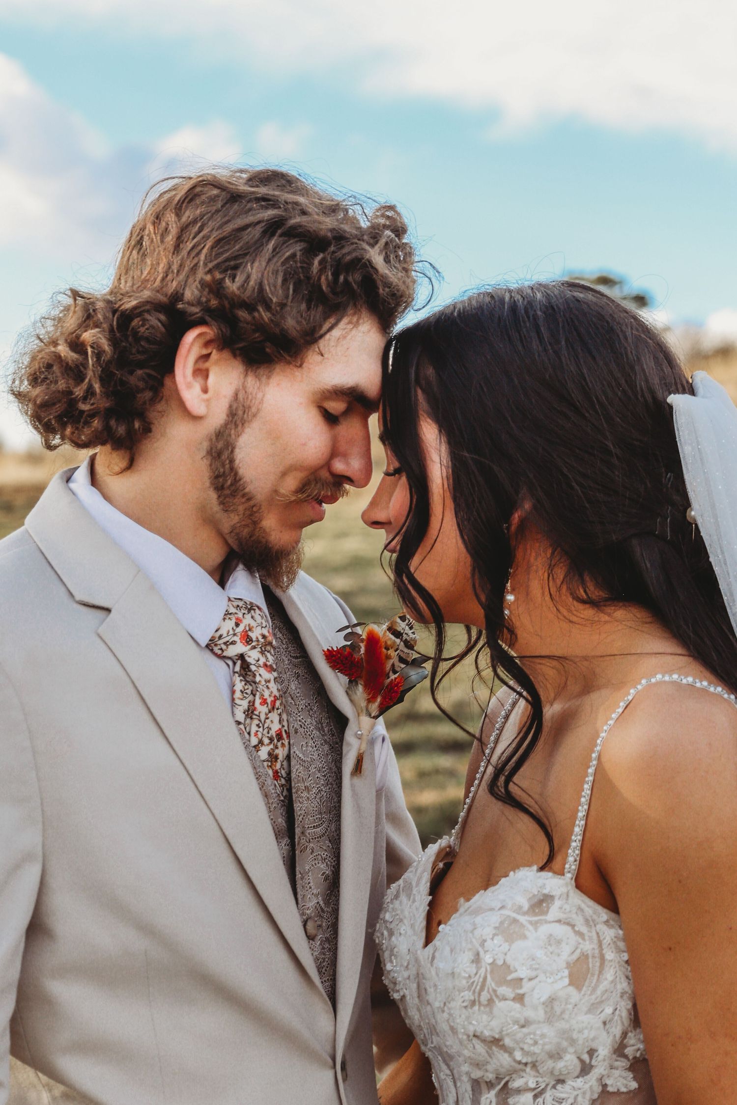 Close-up of bride and groom touching foreheads tenderly, groom in grey suit with floral tie and bride in lace wedding gown.