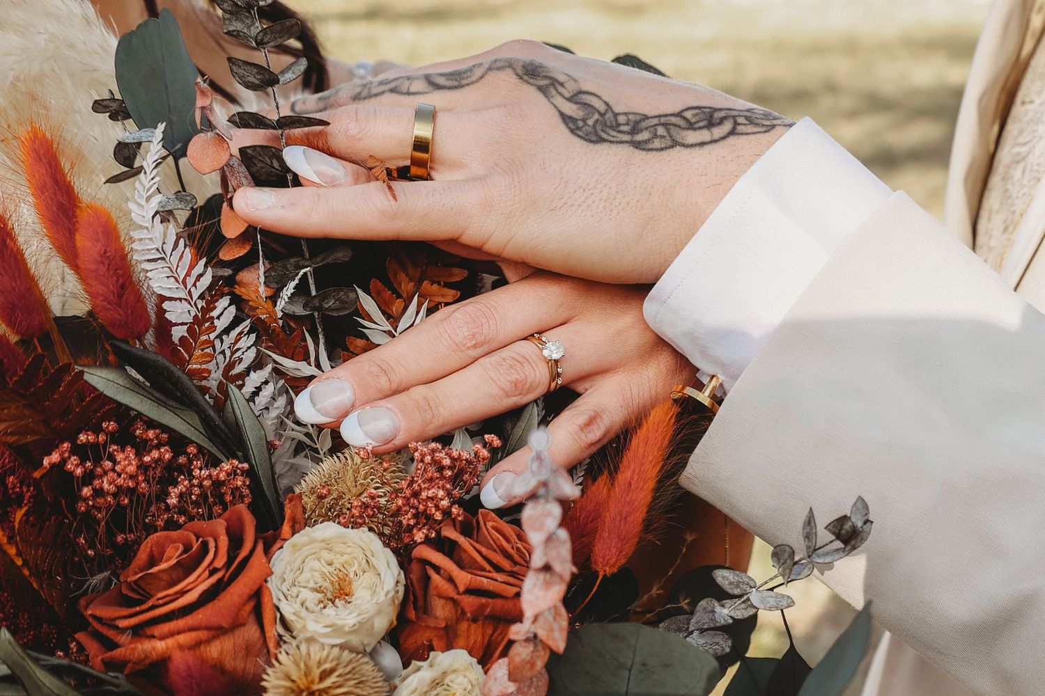 Close-up of newlyweds hands displaying wedding rings resting on a rustic autumn bridal bouquet.
