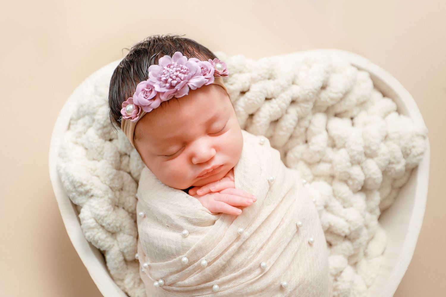 Sleeping newborn baby swaddled in cream wrap with pink floral headband resting in heart-shaped knit basket.