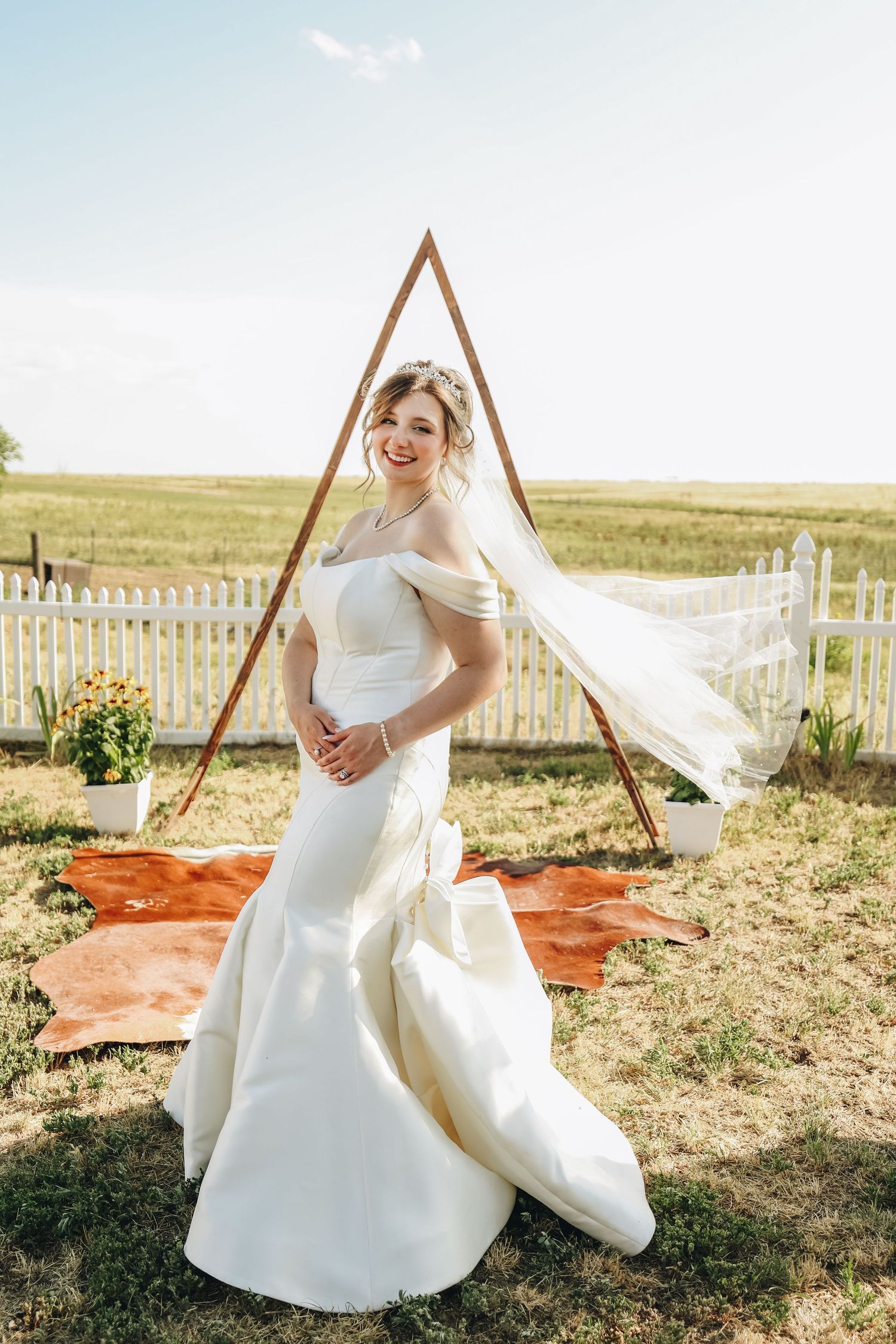 Smiling bride in off-shoulder white mermaid gown stands before triangular wooden arch at outdoor farm wedding.