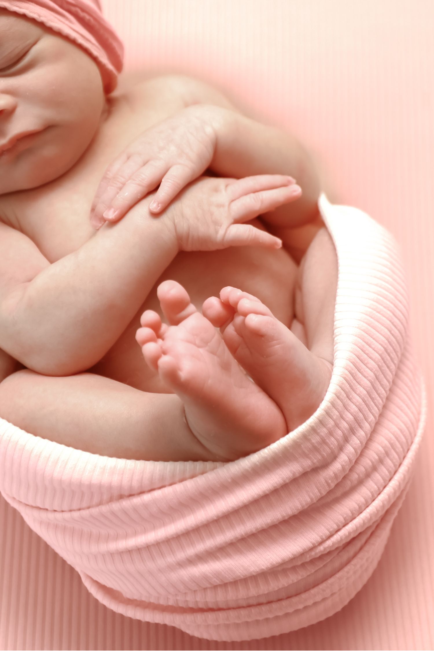 Newborn baby girl sleeping curled up in pink swaddle blanket with matching hat, tiny feet and hands visible.