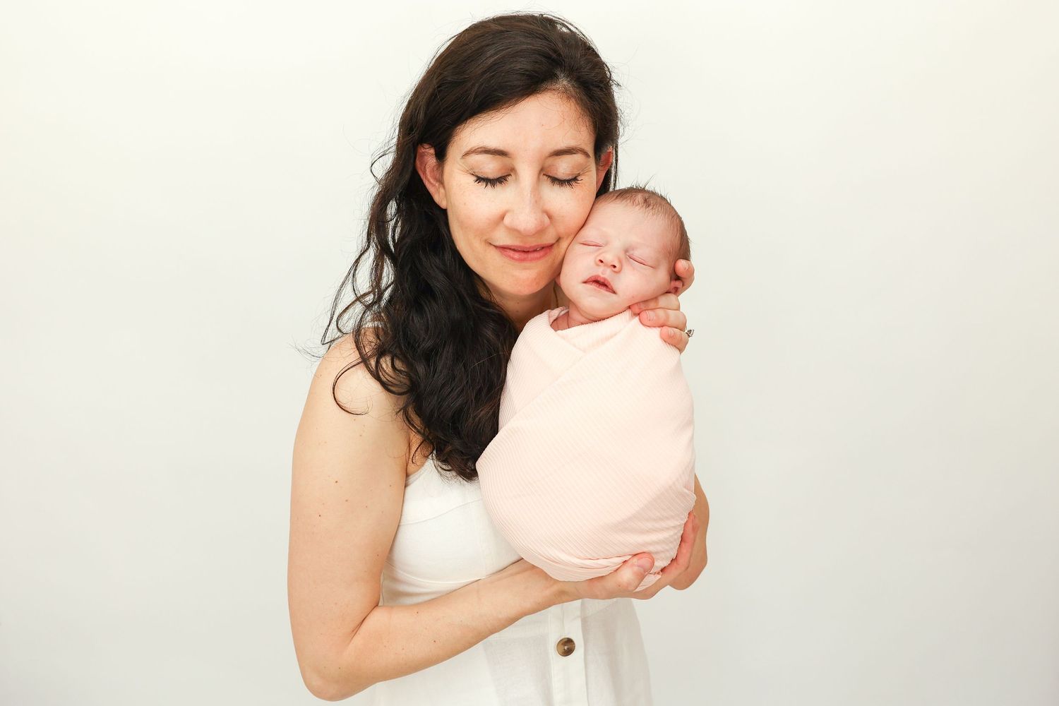 Happy mother in white dress gently holding swaddled newborn baby against white background.