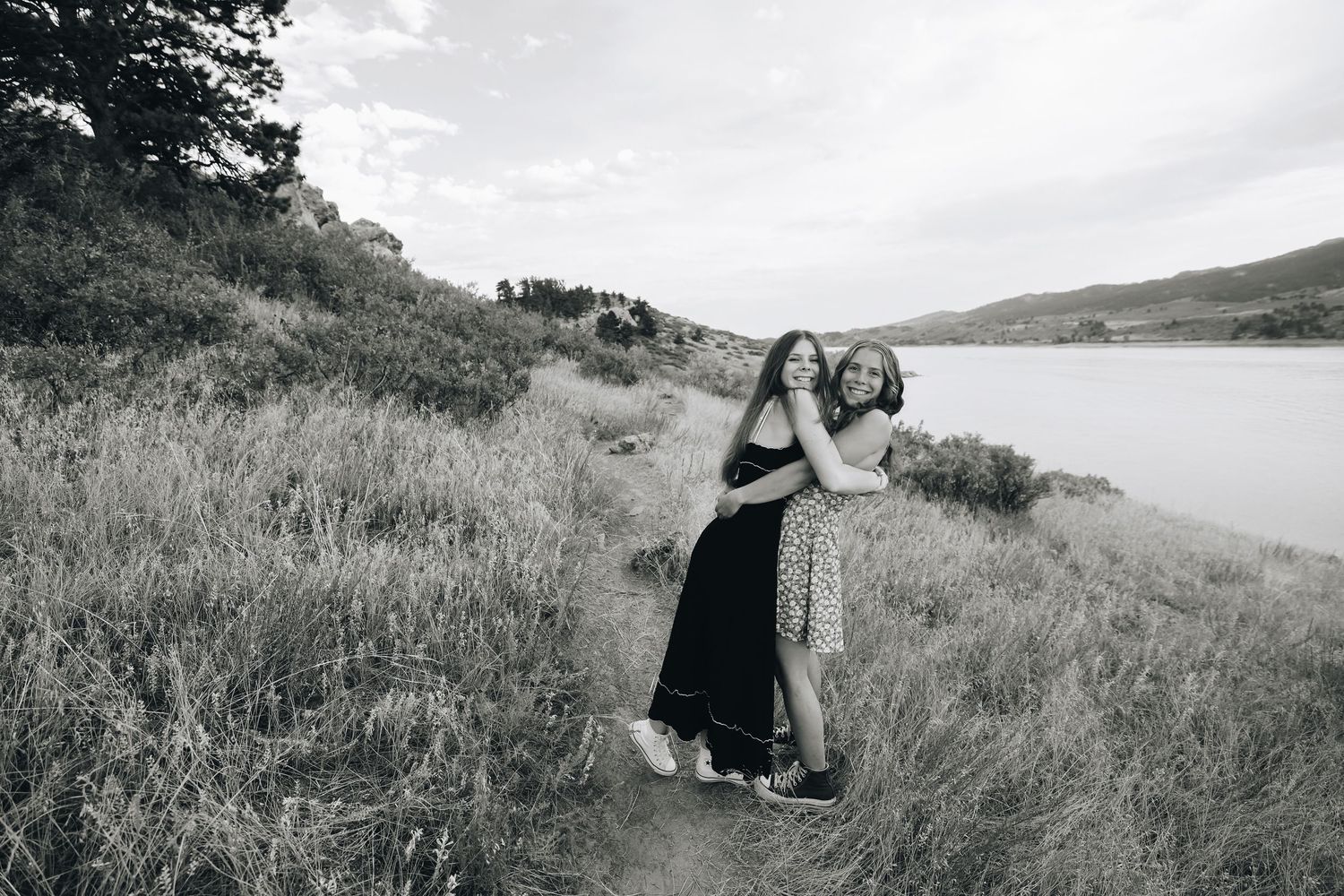 Two young women embracing on a hillside path near a lake in black and white photo.