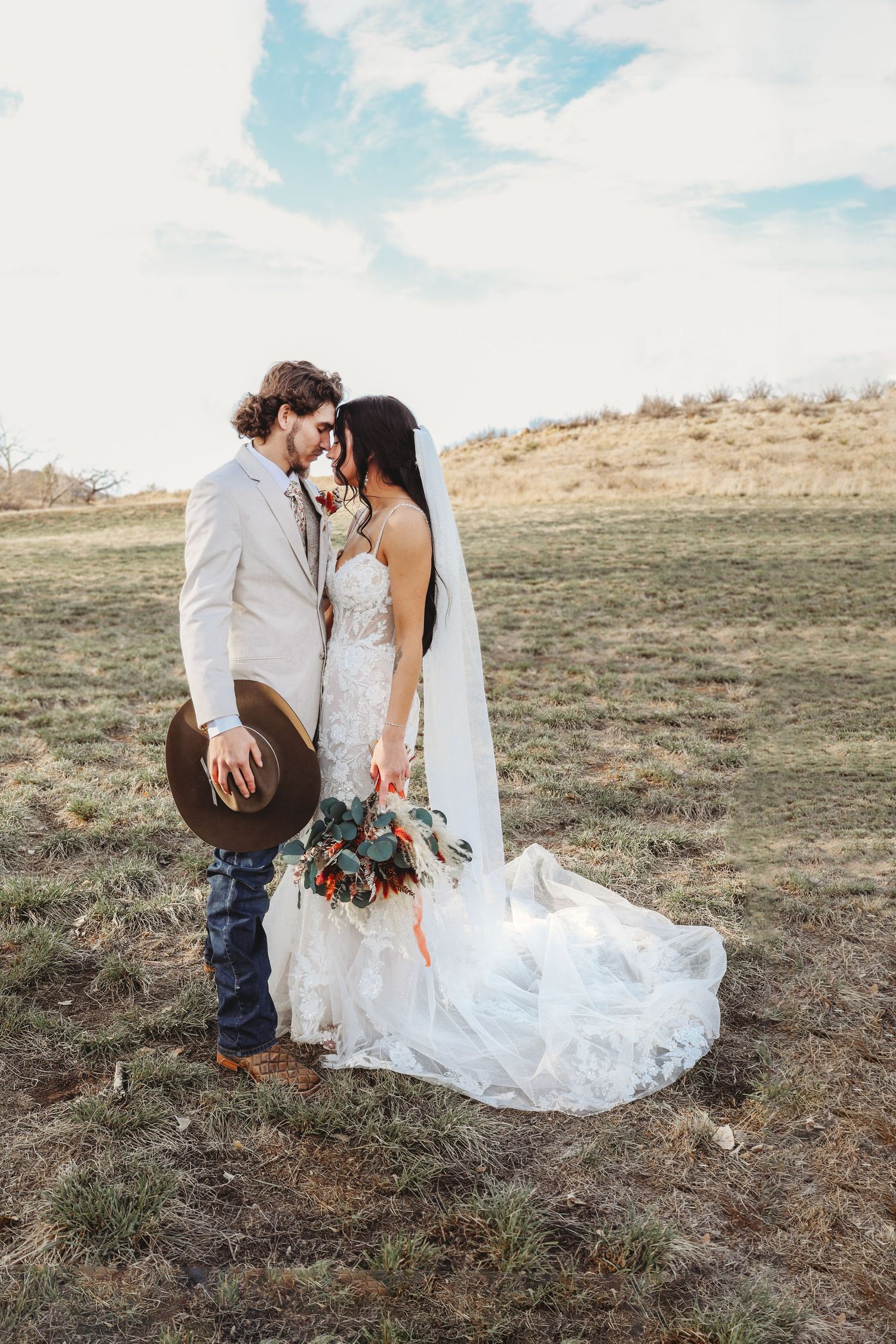 Bride and groom share a romantic moment in an open field, groom holding a cowboy hat and bride carrying a boho bouquet.