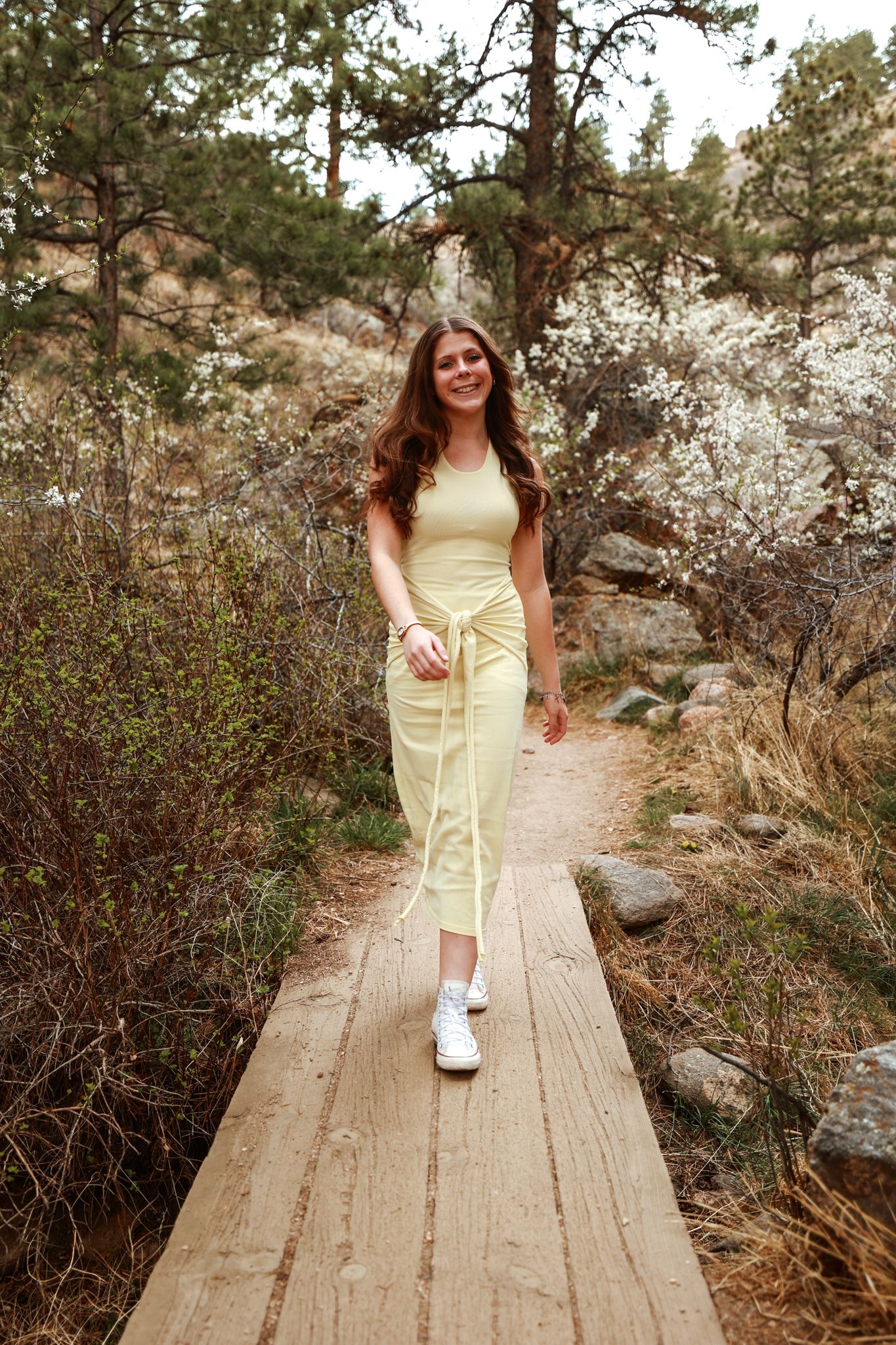Young woman in yellow midi dress walking on wooden path through scenic mountain forest trail.