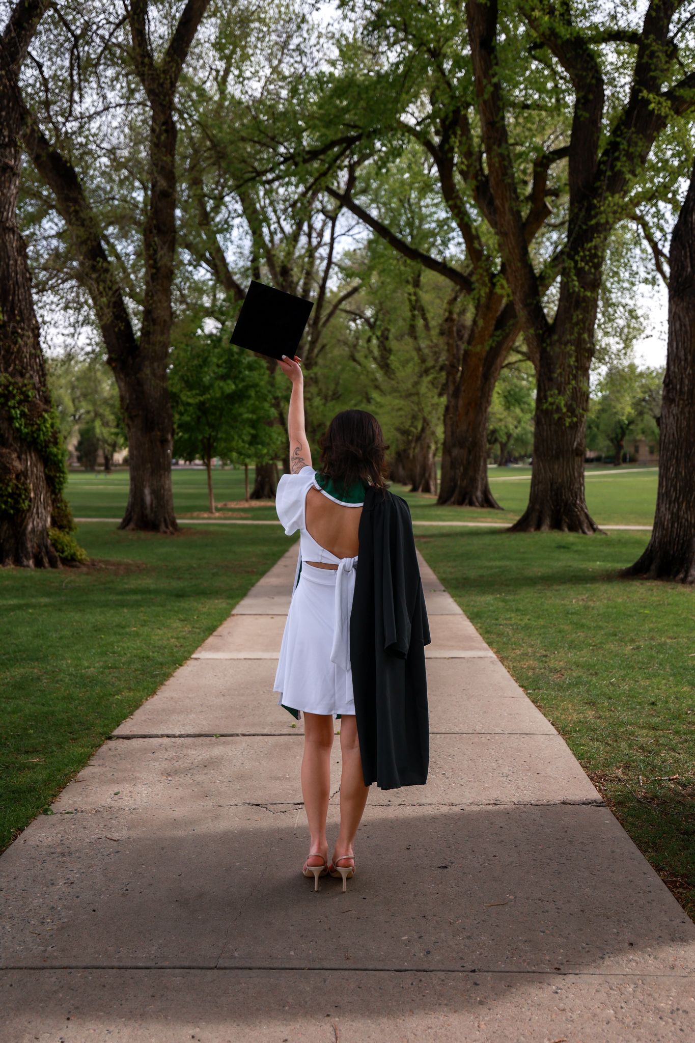 A graduate in white dress holds cap aloft on tree-lined campus walkway, celebrating academic achievement.