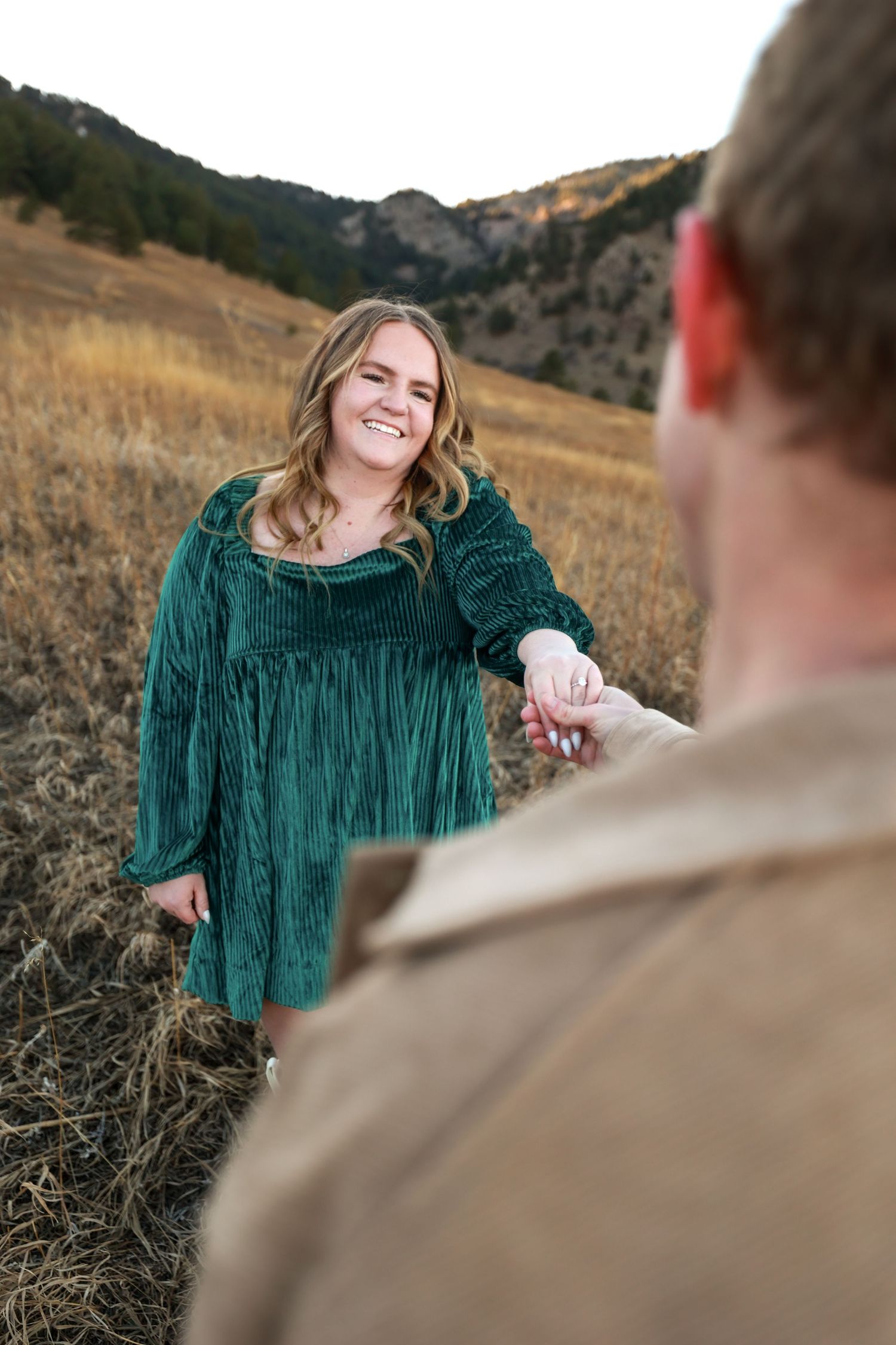 Smiling woman in green velvet dress holds mans hand in golden mountain field during outdoor engagement session.
