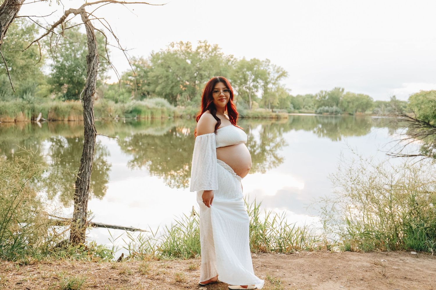 Pregnant woman in flowing white dress holding belly posing near calm reflective lake outdoors.