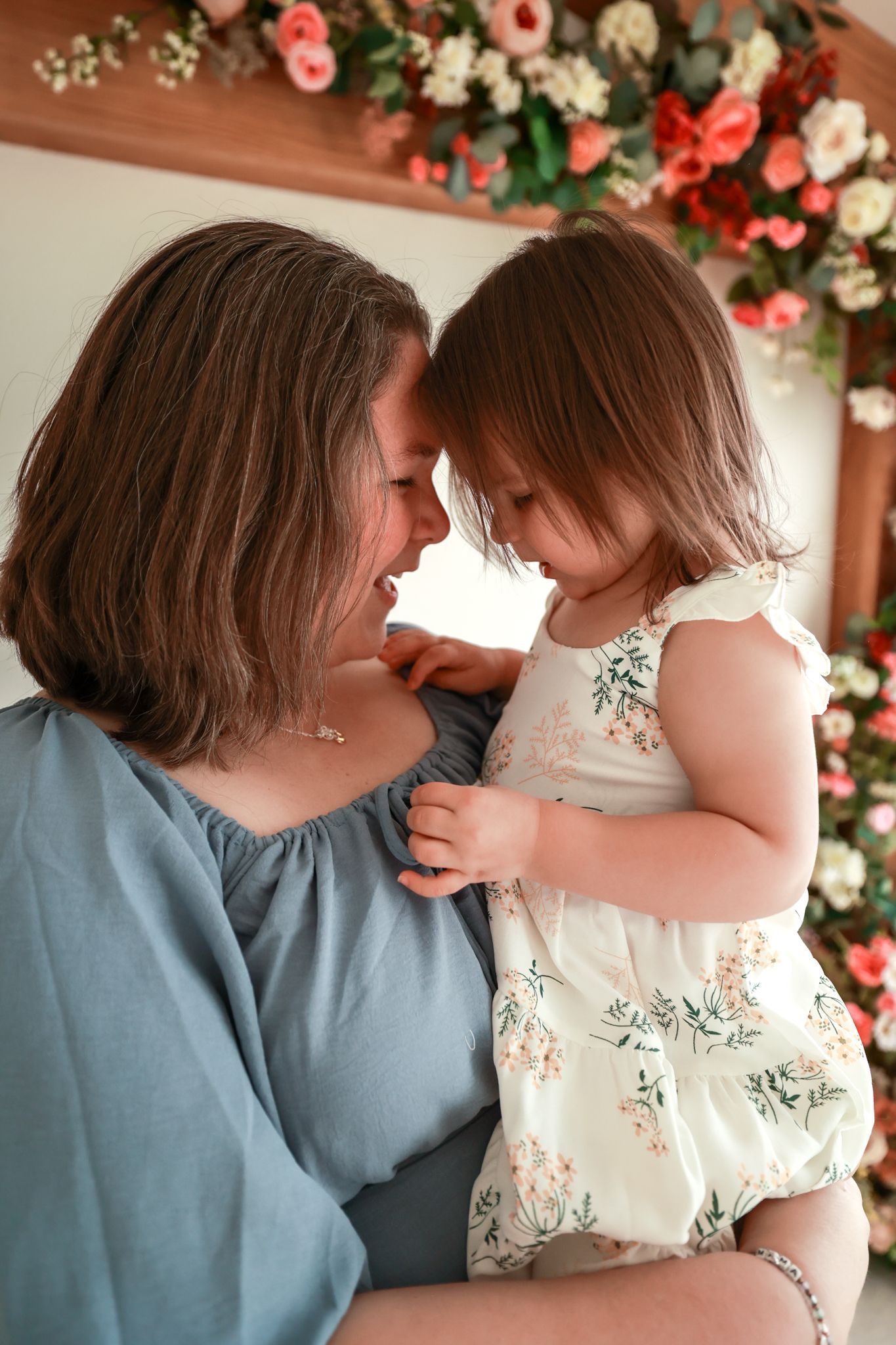 A mother and toddler daughter share a tender forehead touch, surrounded by pink and white flowers.