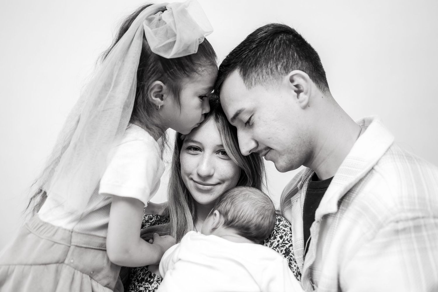 Black and white family portrait of parents tenderly holding newborn baby with young daughter kissing mothers cheek.