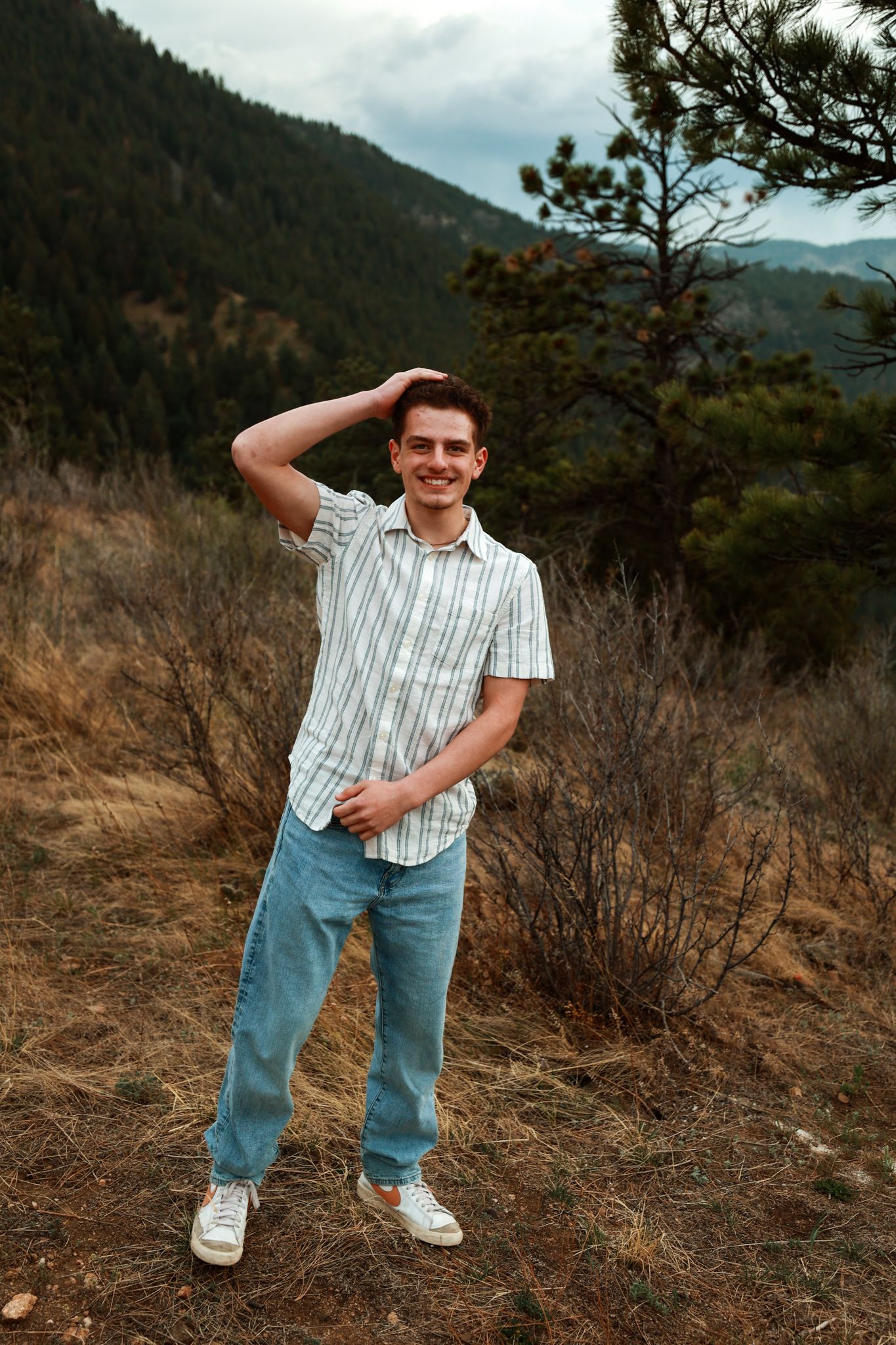 Young man in striped shirt and jeans smiling outdoors in a mountain forest landscape.