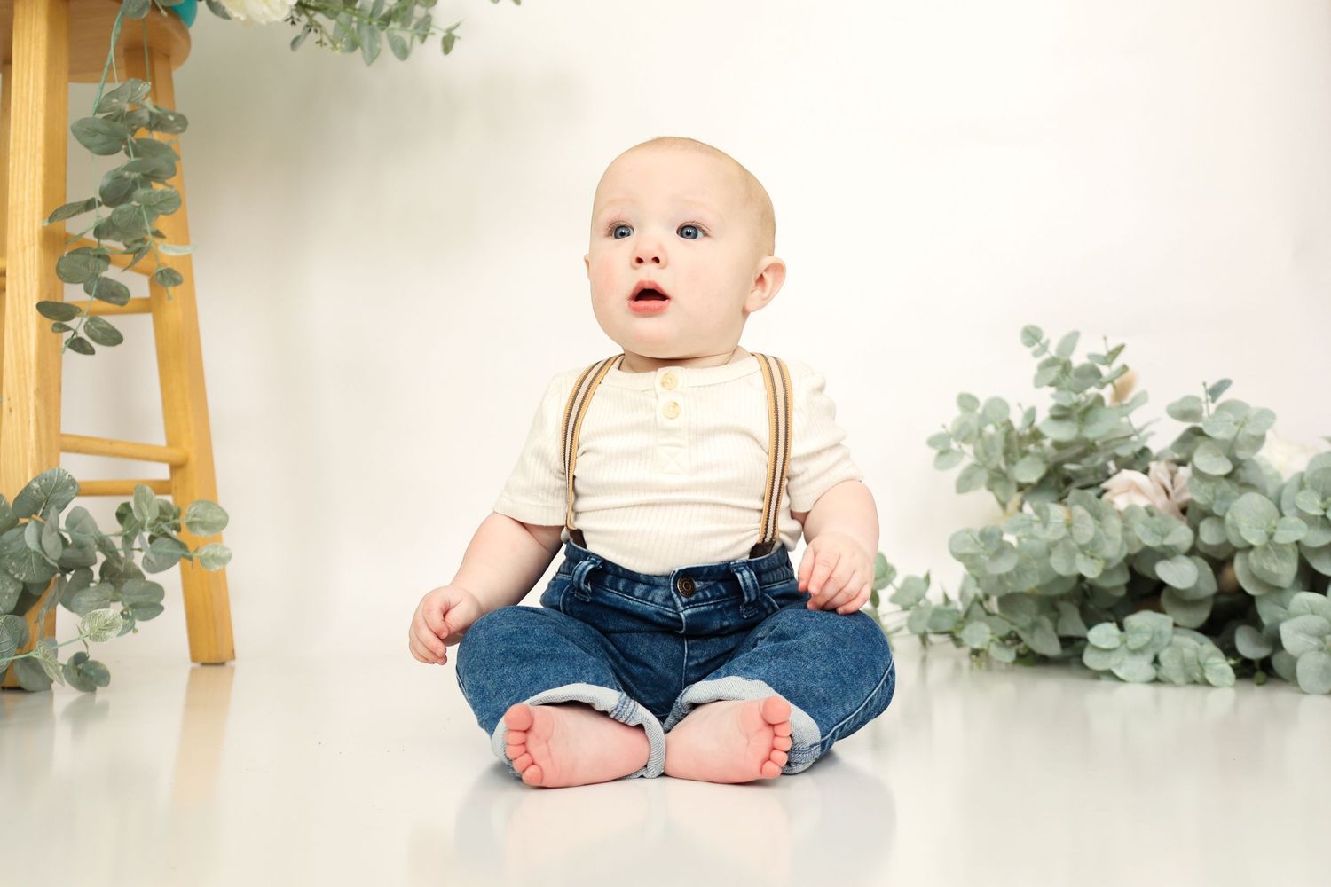 Adorable baby boy in suspenders and jeans sitting on a white studio floor surrounded by eucalyptus decor.
