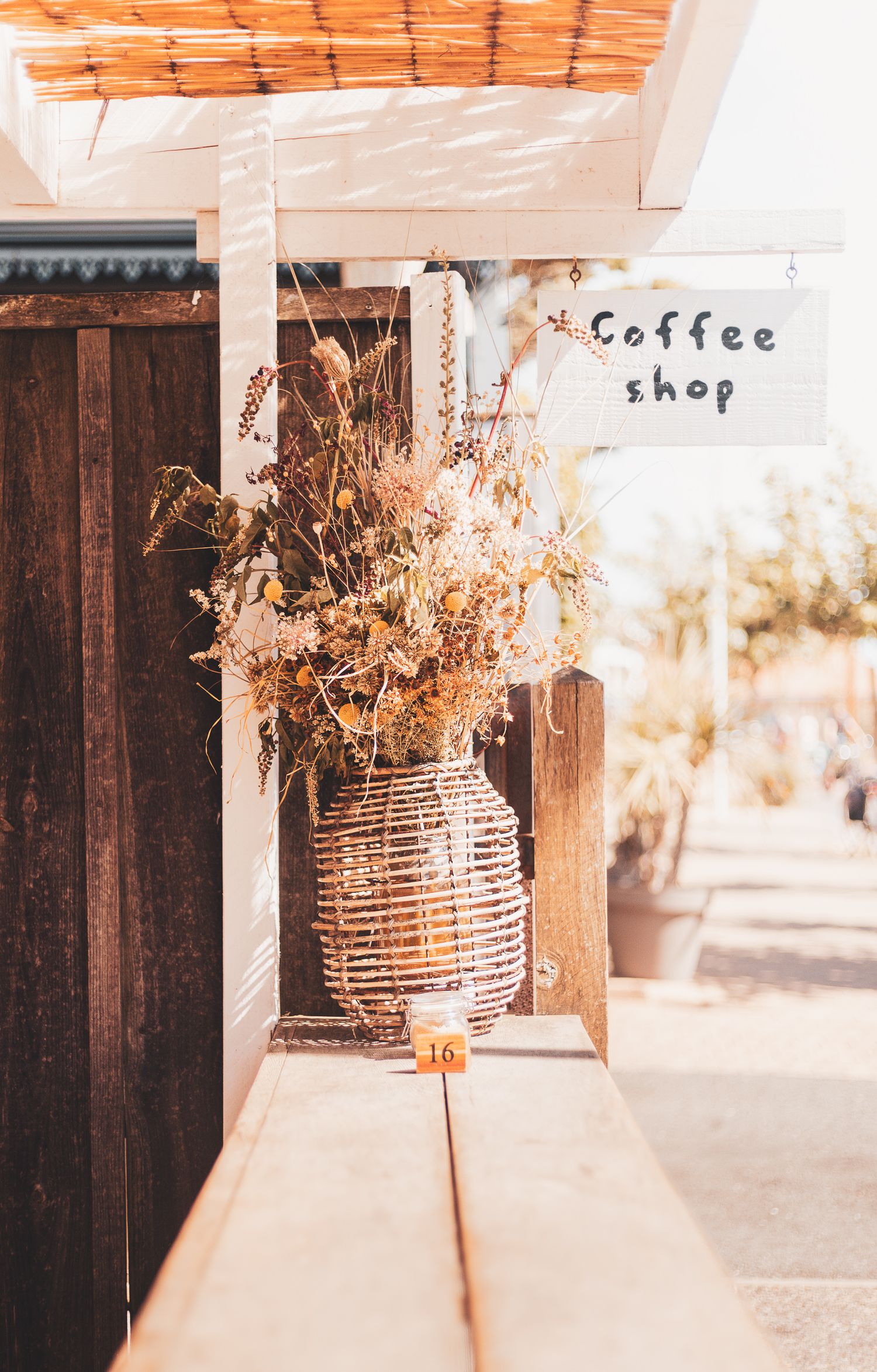 Une entrée de café rustique avec des fleurs séchées dans un panier tissé et un simple panneau manuscrit sur le mur.