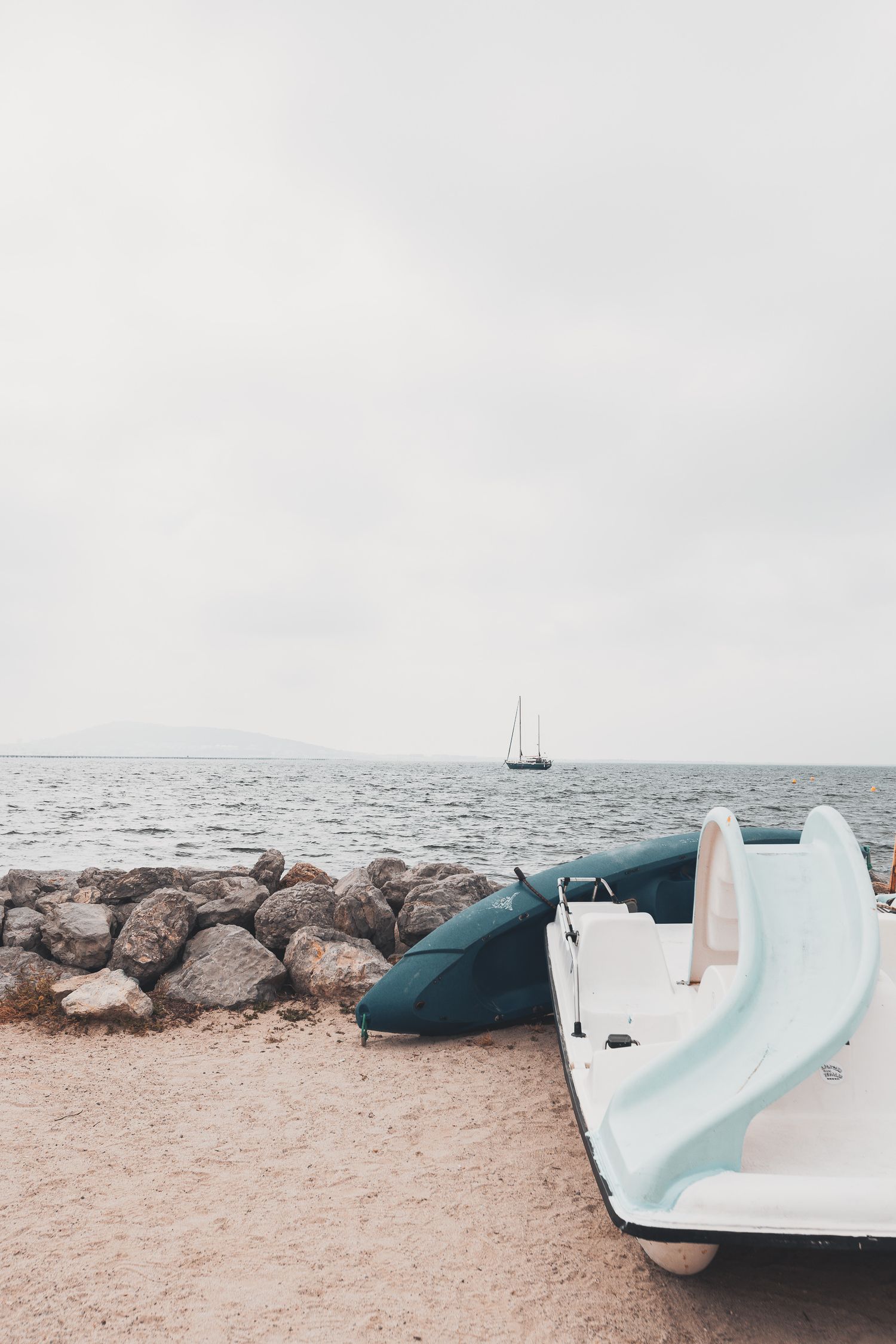 Petits bateaux reposant sur une plage rocheuse avec un voilier visible dans des eaux brumeuses lointaines.