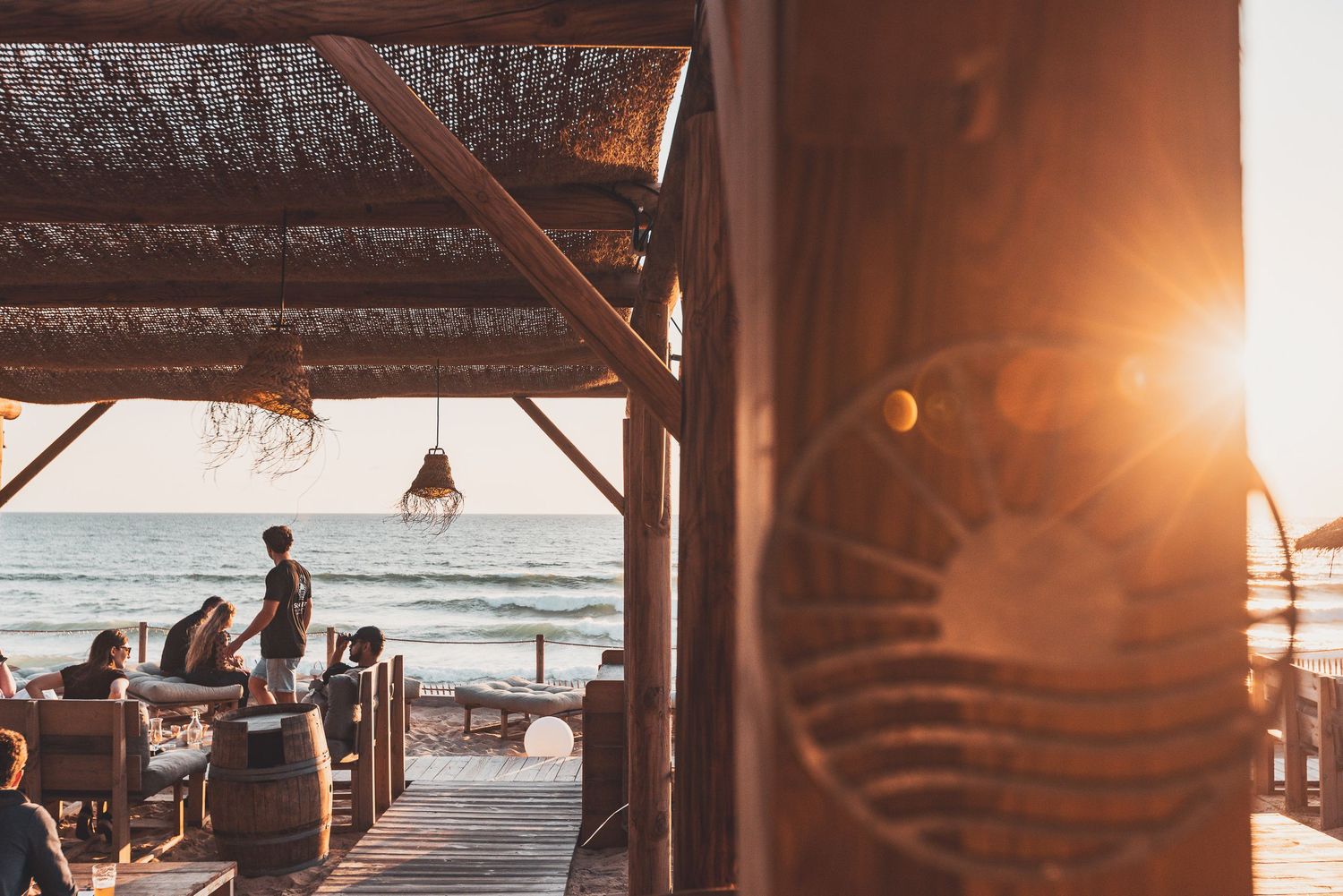 A beachside wooden pavilion at sunset with people silhouetted against the golden light over the ocean.