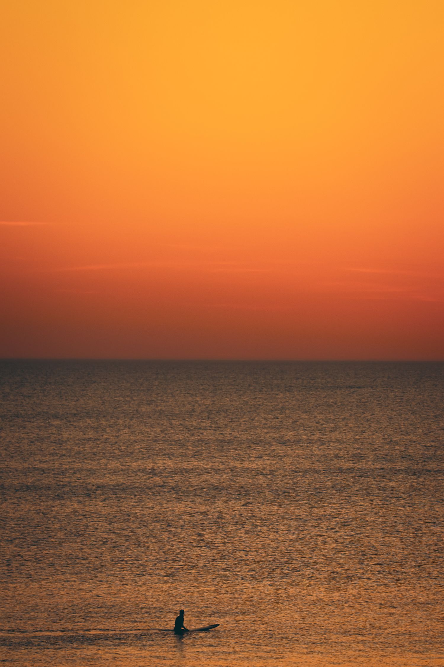 Un surfeur solile silhouette contre un coucher de soleil orange vibrant sur des eaux océaniques calmes crée une scène côtière sereine.