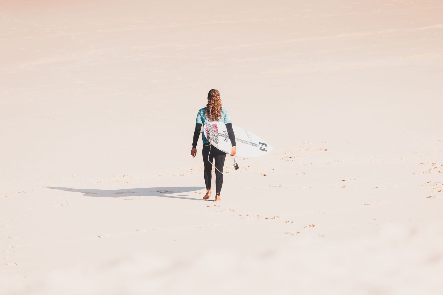 Personne portant une planche de surf marchant sur une plage de sable blanc immaculée.