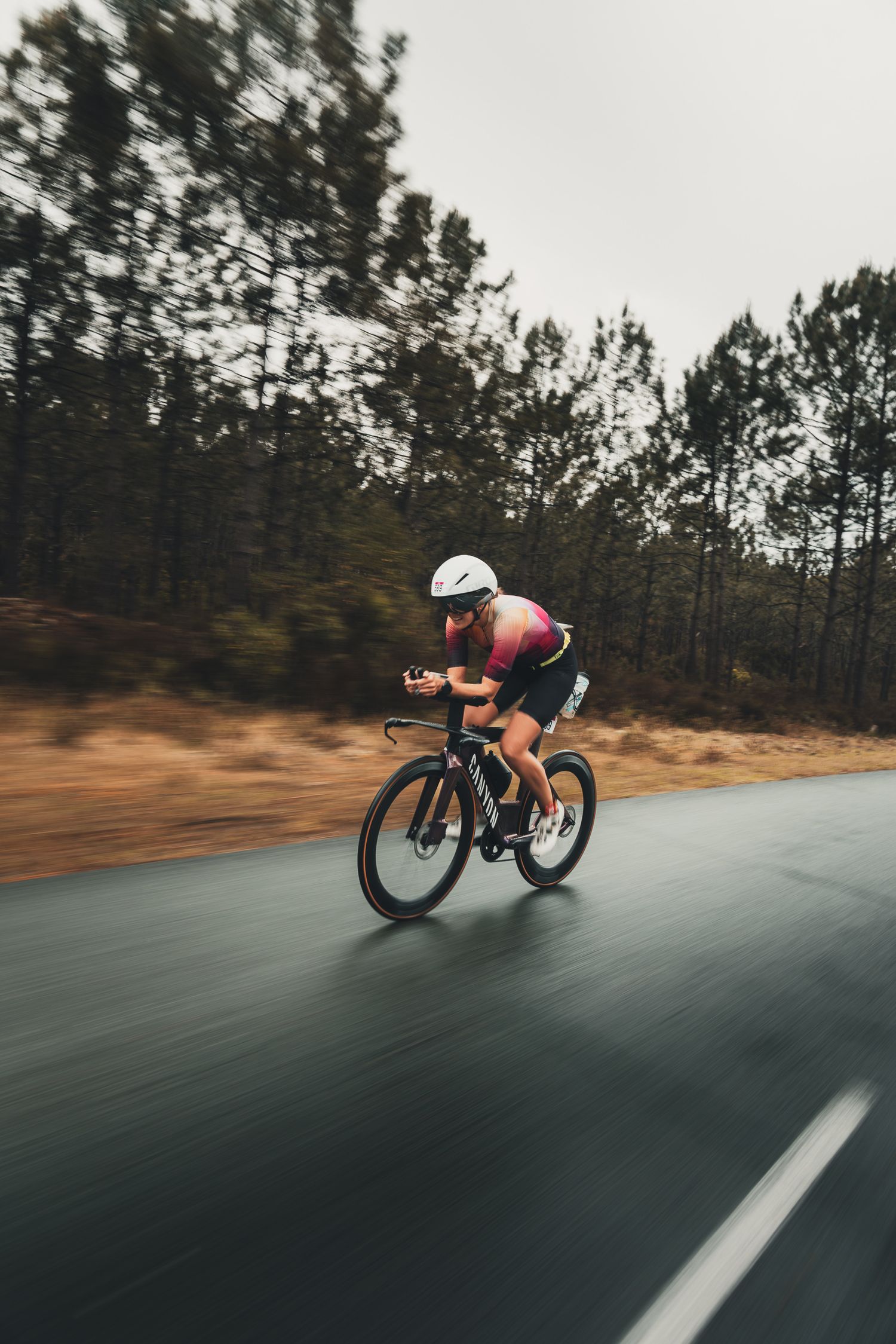 Cyclist in aerodynamic position racing at high speed on a wet road surrounded by pine trees, wearing a white helmet and pink jersey.