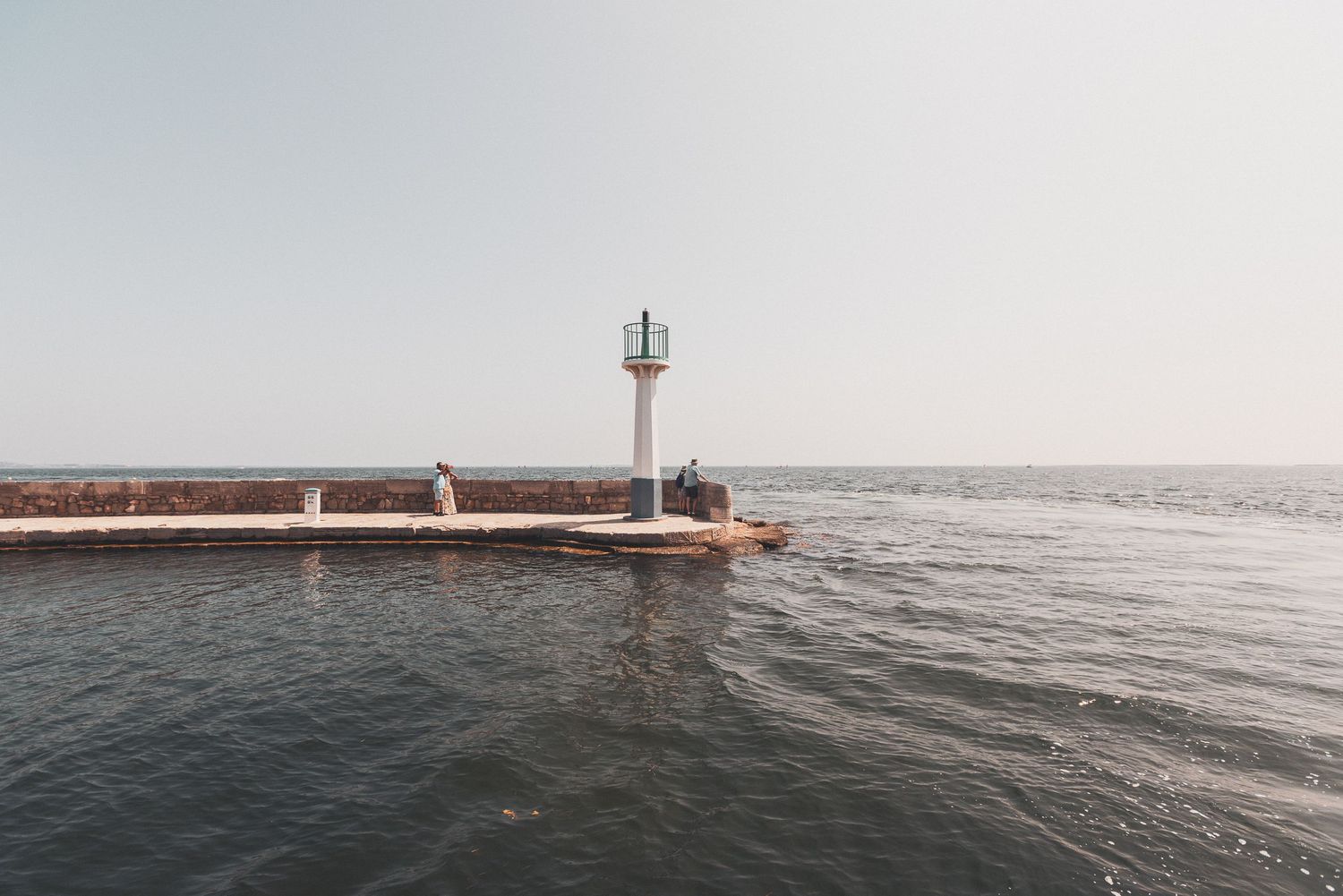A lighthouse stands at the end of a concrete pier extending into misty ocean waters.