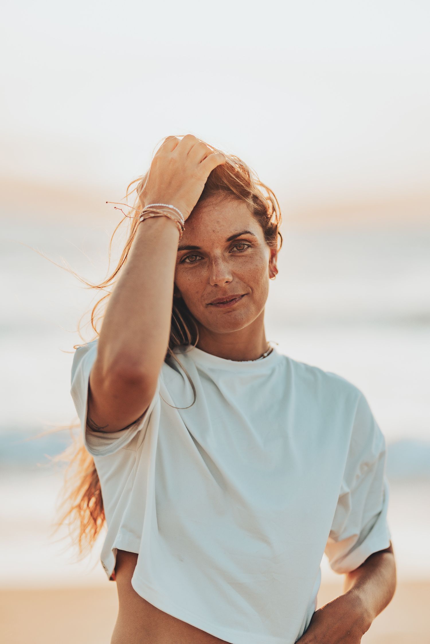 Personne en t-shirt blanc avec des cheveux balayés par le vent posant sur la plage au coucher du soleil