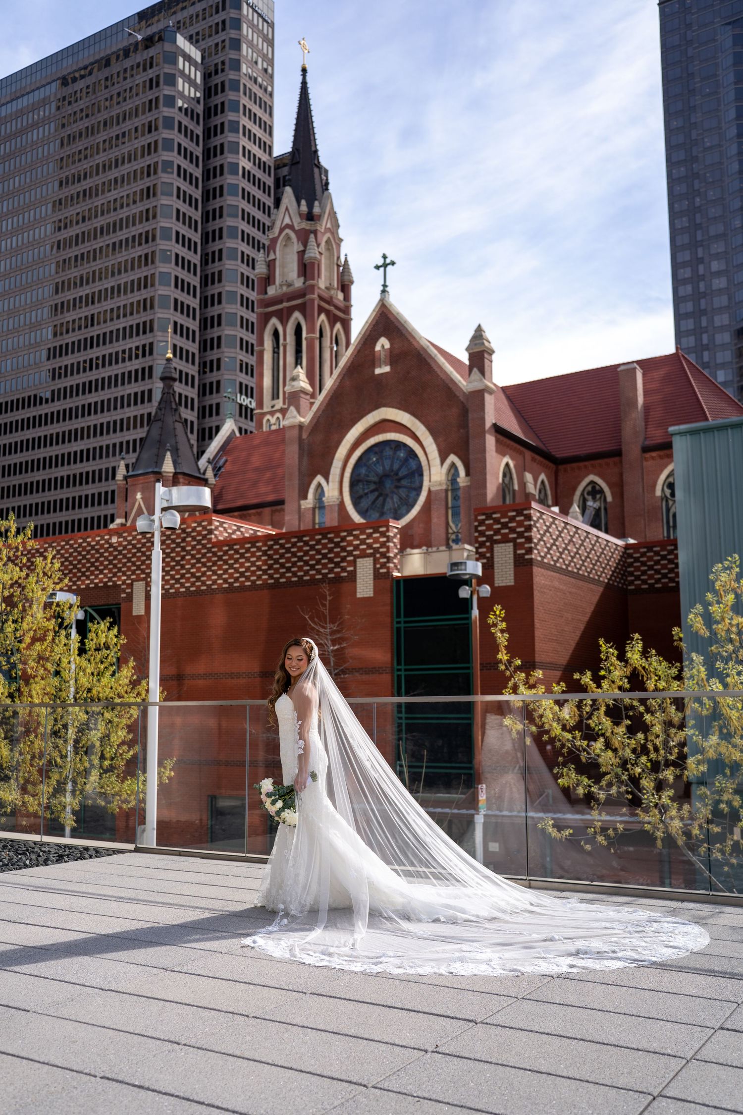 Bride in white gown and long veil poses before a red brick Gothic church in an urban setting with modern skyscrapers.