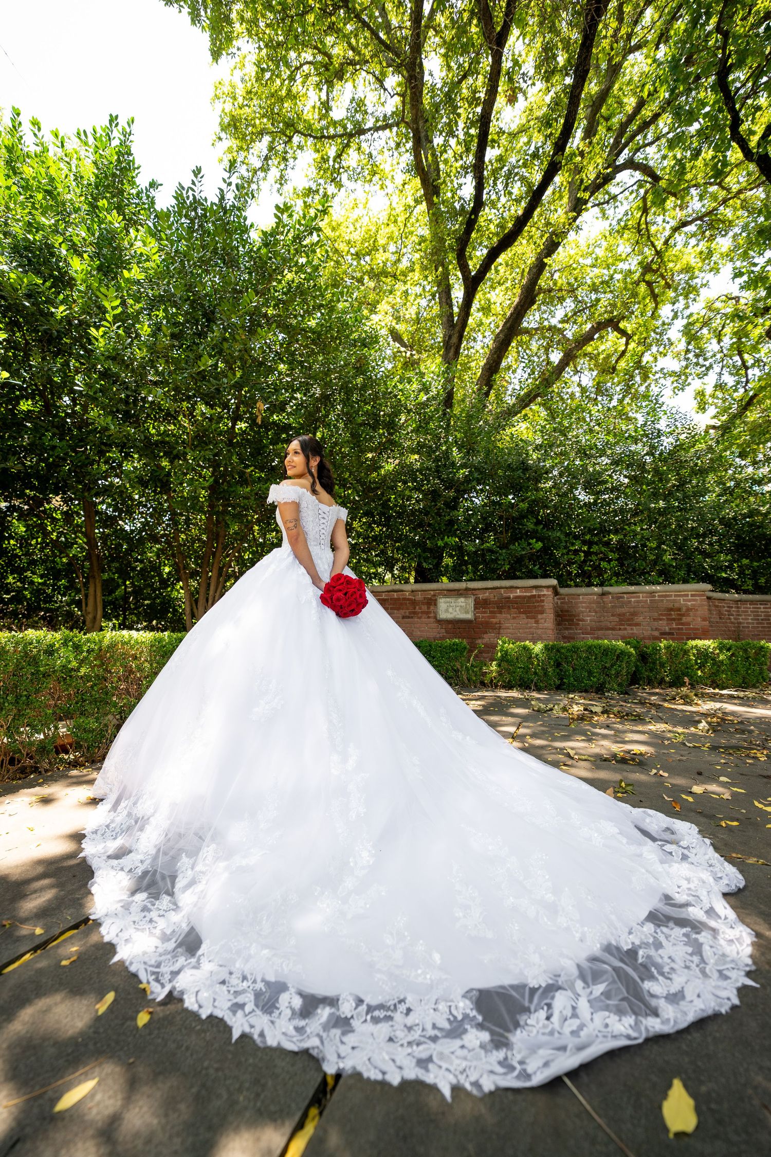 A bride in a flowing white ball gown with a dramatic train poses in a garden setting with brick walls and trees.