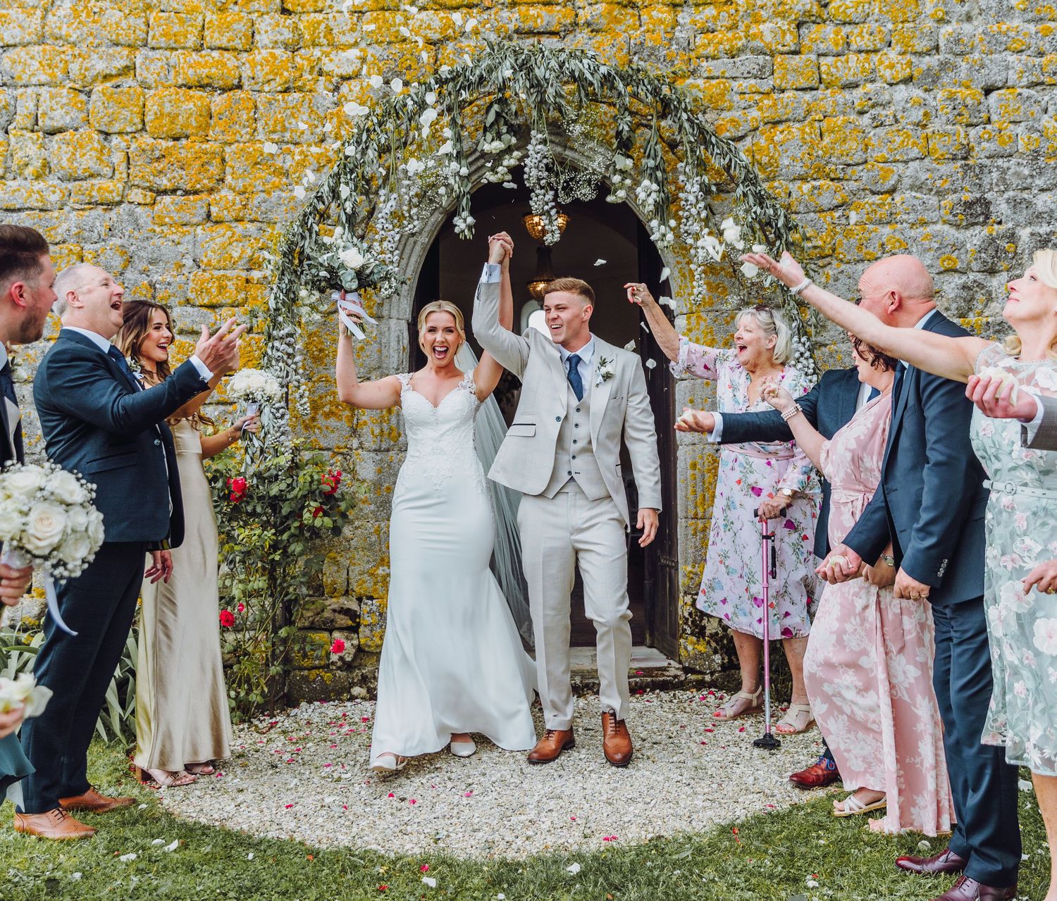Wedding party celebrates with confetti toss along rustic stone wall during outdoor garden ceremony.