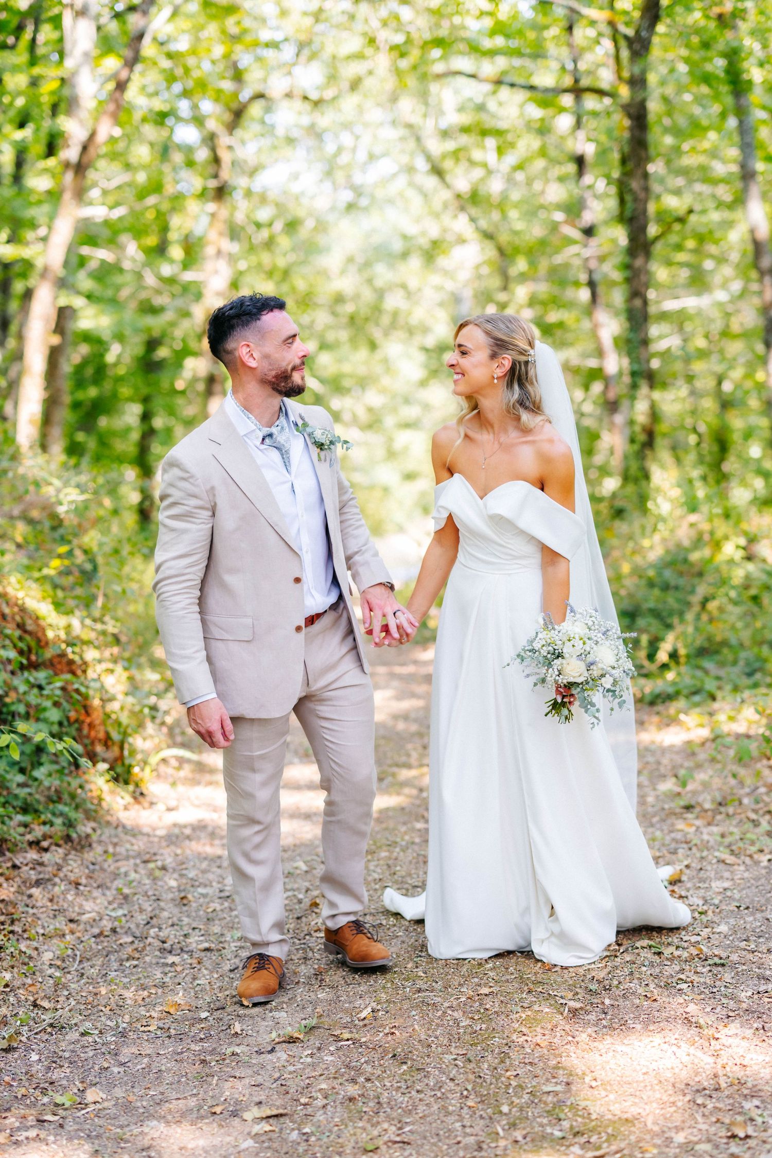 Smiling bride in an off-shoulder white gown and groom in a beige suit hold hands on a sunlit forest path during their wedding photoshoot.