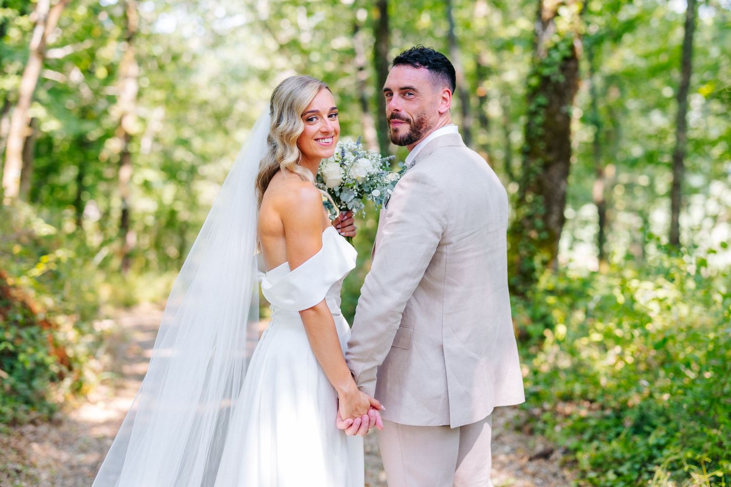 Bride in white off-shoulder gown and groom in beige suit hold hands in a lush green forest wedding photo.