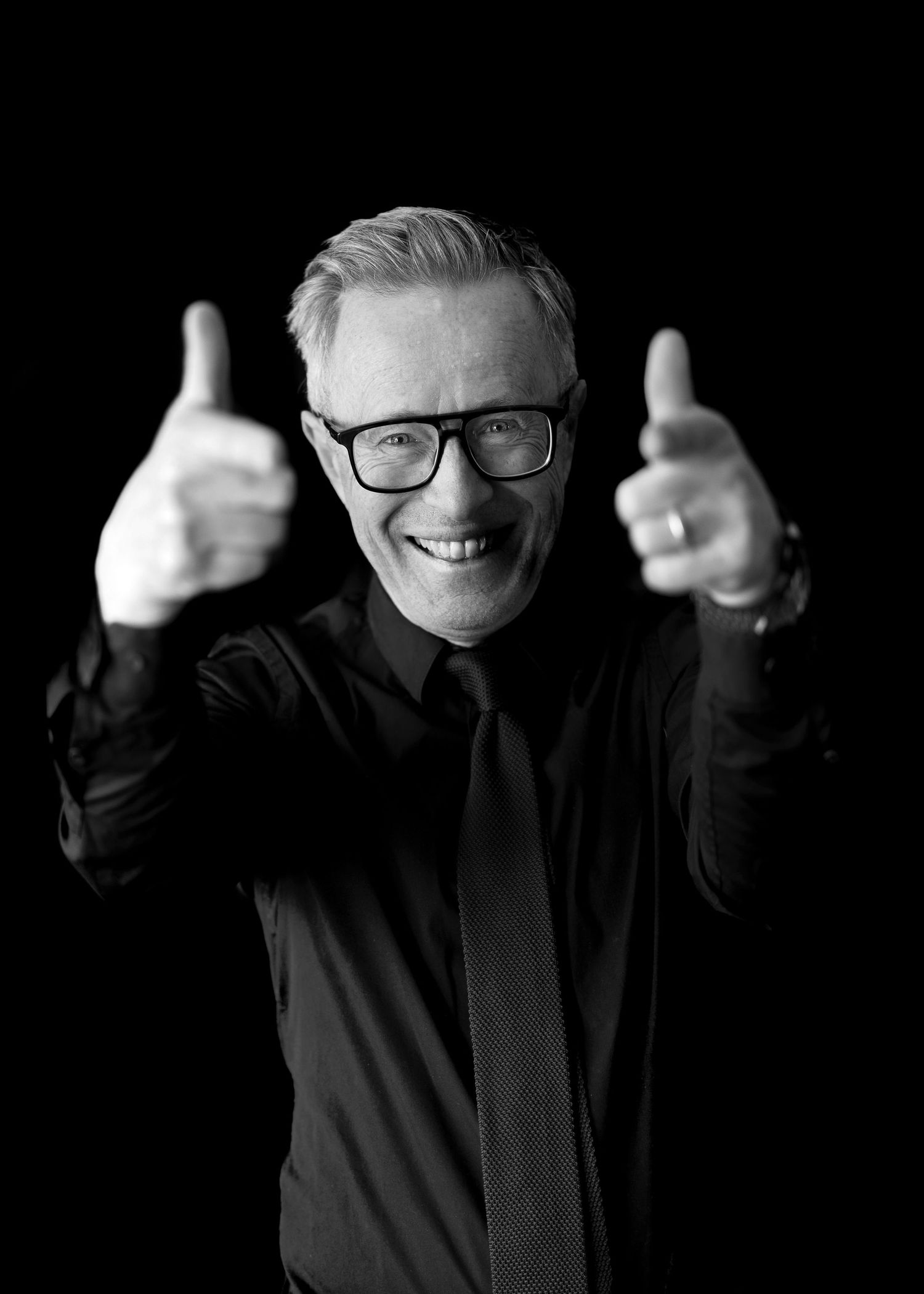 Smiling man in black shirt and tie giving two thumbs up, wearing glasses, black and white portrait photo.