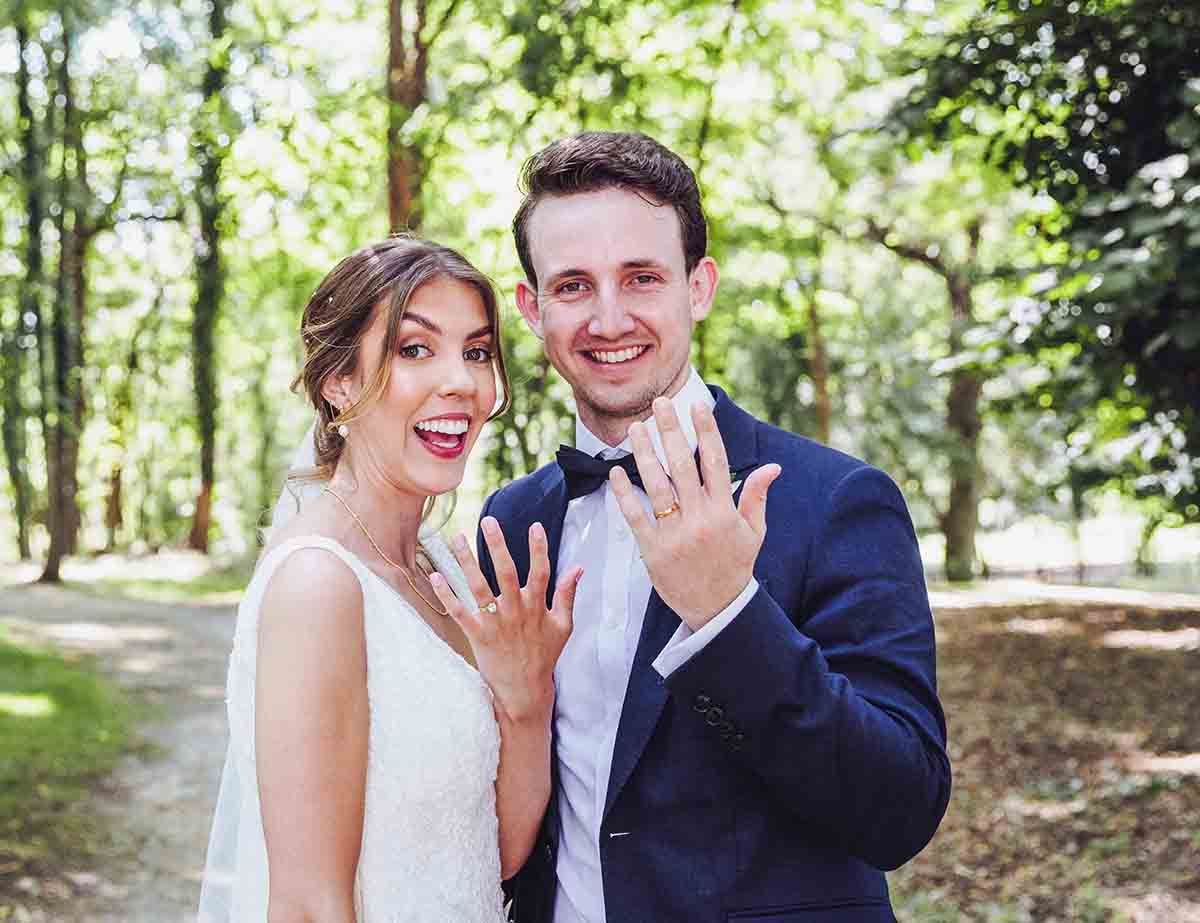 A newly engaged couple showing off their rings while standing together in a sunlit wooded park setting.