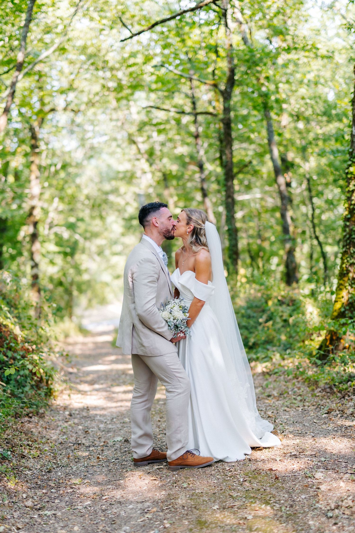A bride and groom share a romantic kiss on a sunlit forest path, surrounded by lush green trees on their wedding day.