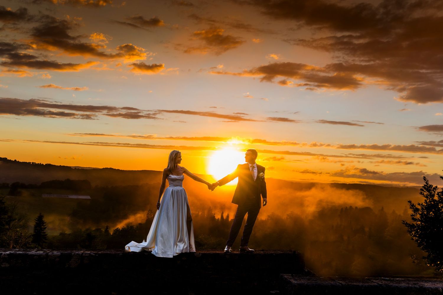 Bride in a flowing white gown and groom in a dark suit hold hands, silhouetted against a golden sunset over rolling hills.