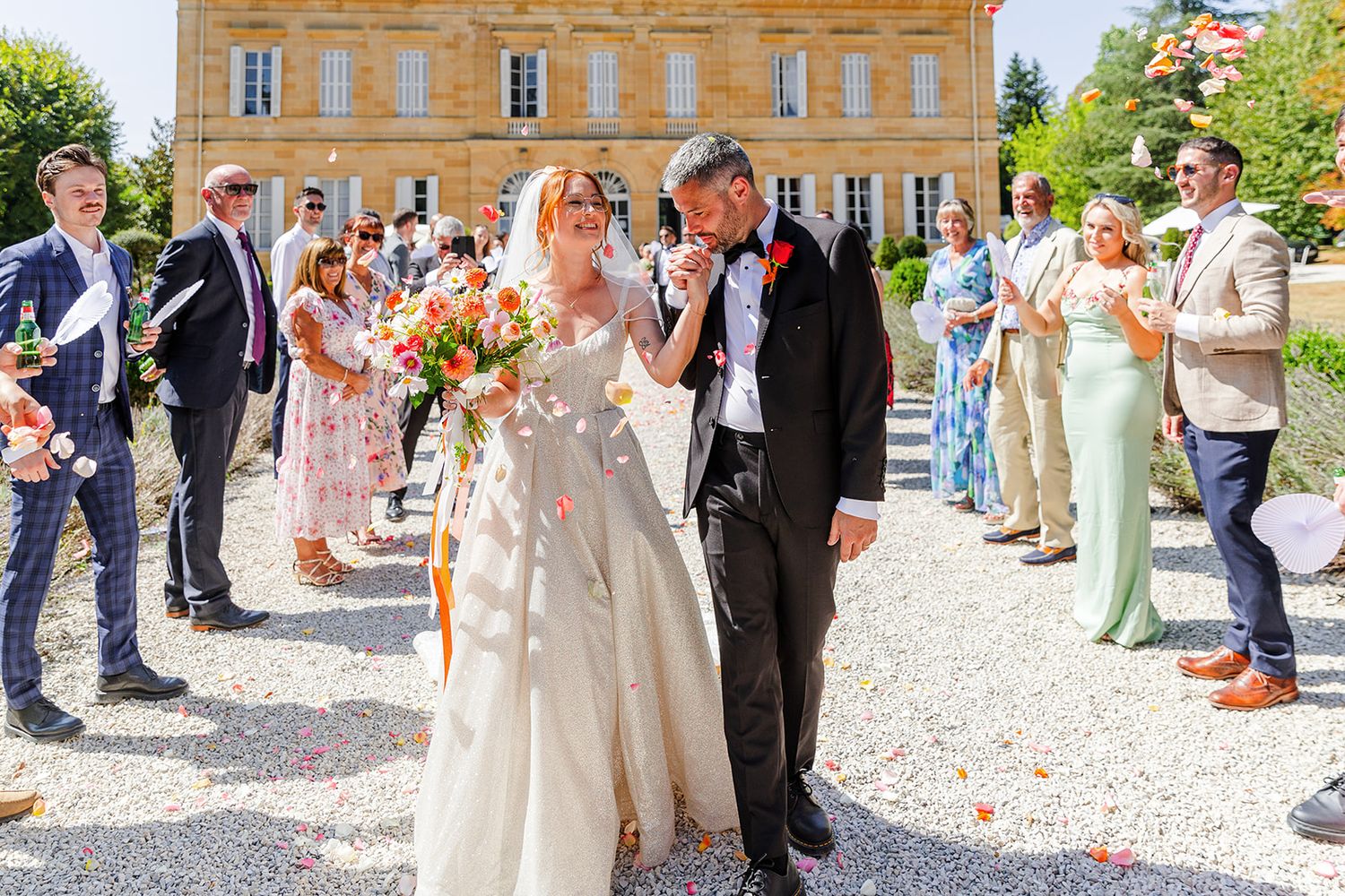 Bride and groom walk down outdoor aisle as guests toss flower petals at elegant French chateau wedding ceremony.