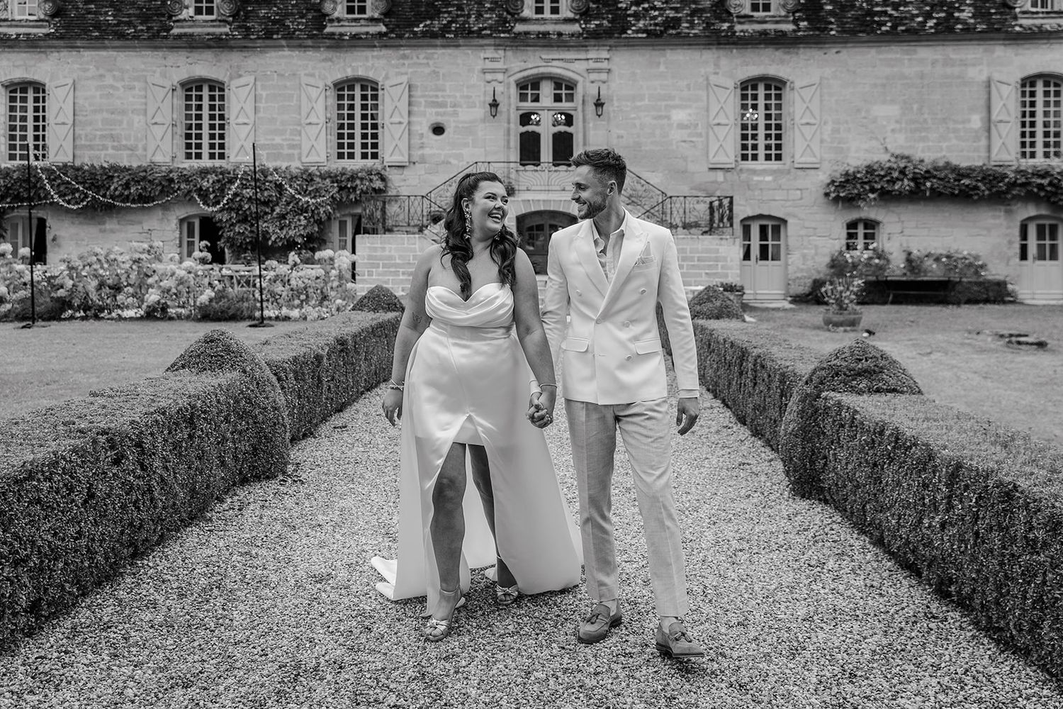 A couple in white outfits walks together on a gravel path between manicured hedges at an elegant French chateau.