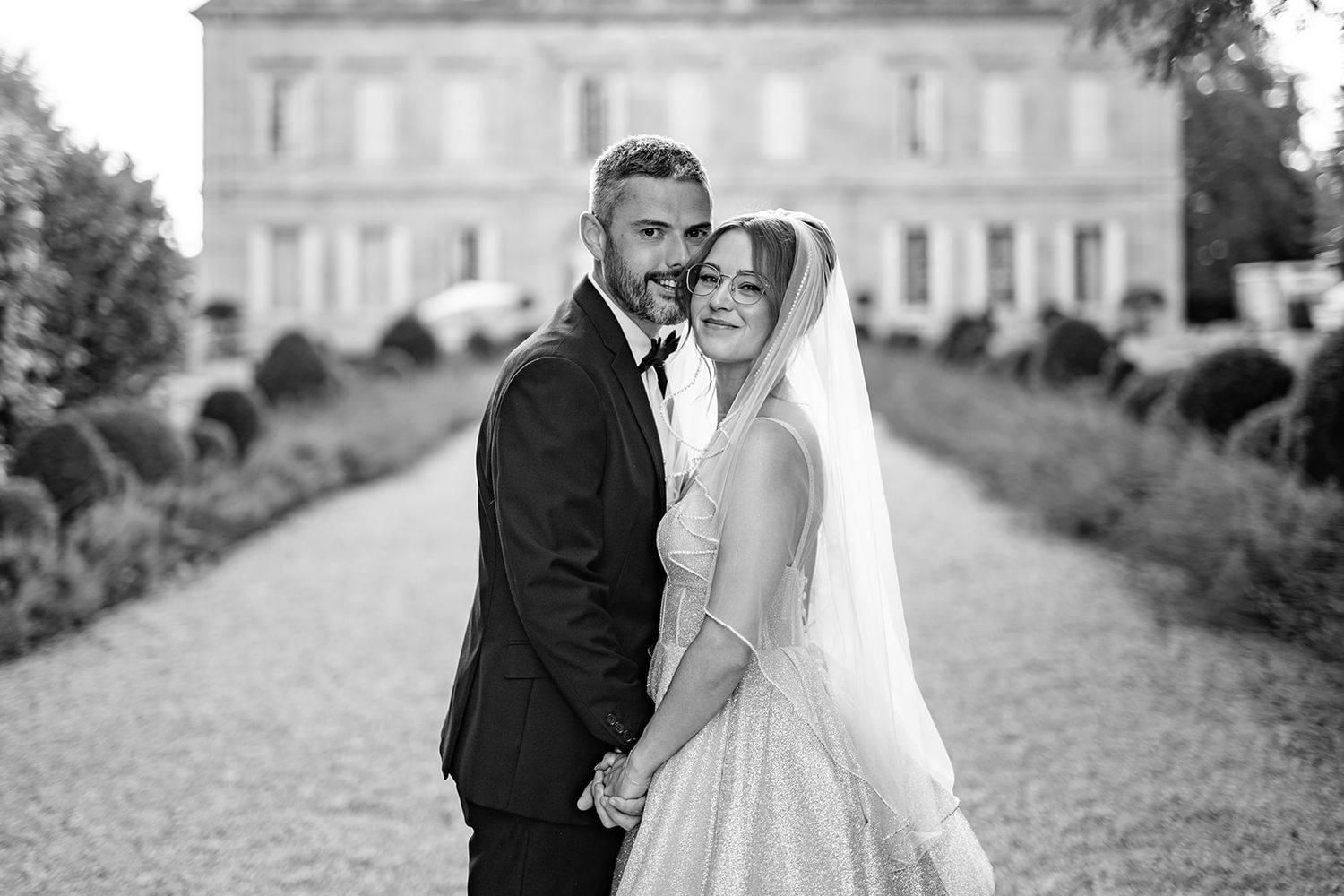 Black and white wedding photo of a bride and groom embracing on a garden path with elegant mansion in background.