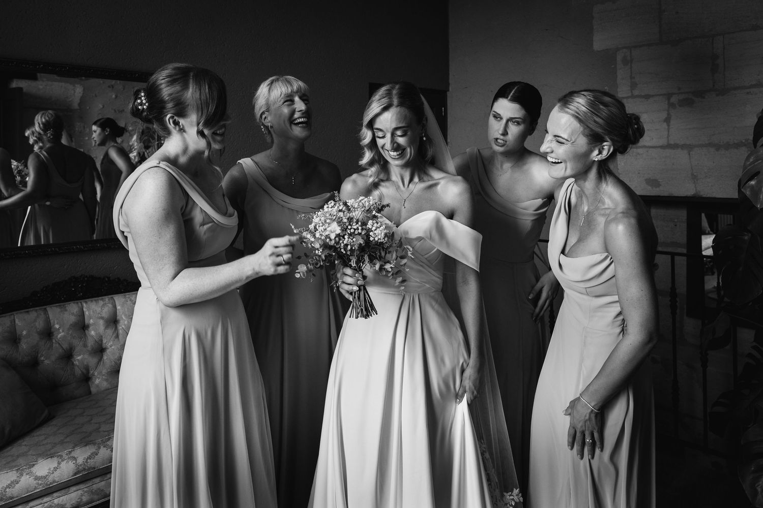 Black and white photo of a bride in white gown holding bouquet surrounded by smiling bridesmaids in elegant dresses.