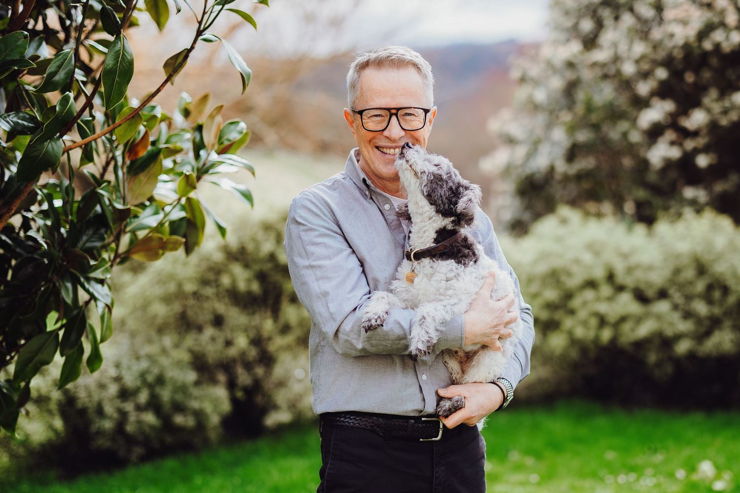 A person in glasses holds a small gray dog while standing in a blooming spring garden with white flowers.