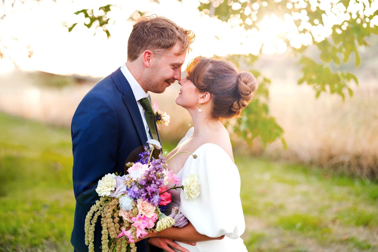 A romantic outdoor wedding moment with soft sunlight filtering through trees and pink floral bouquet details.