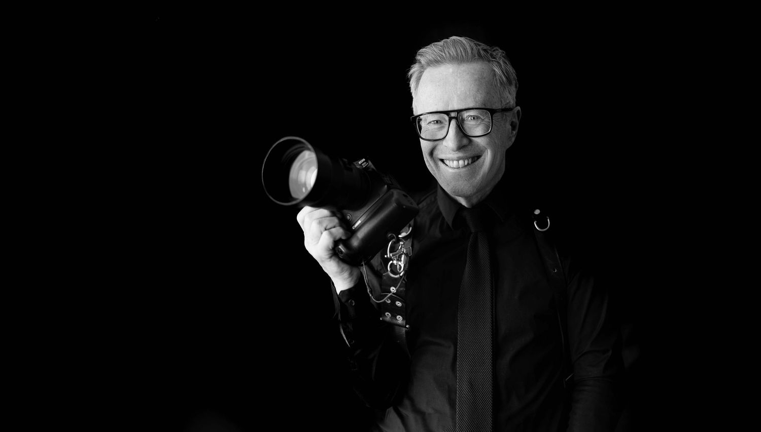 Black and white portrait of a smiling man holding a professional camera, dressed in dark clothing.