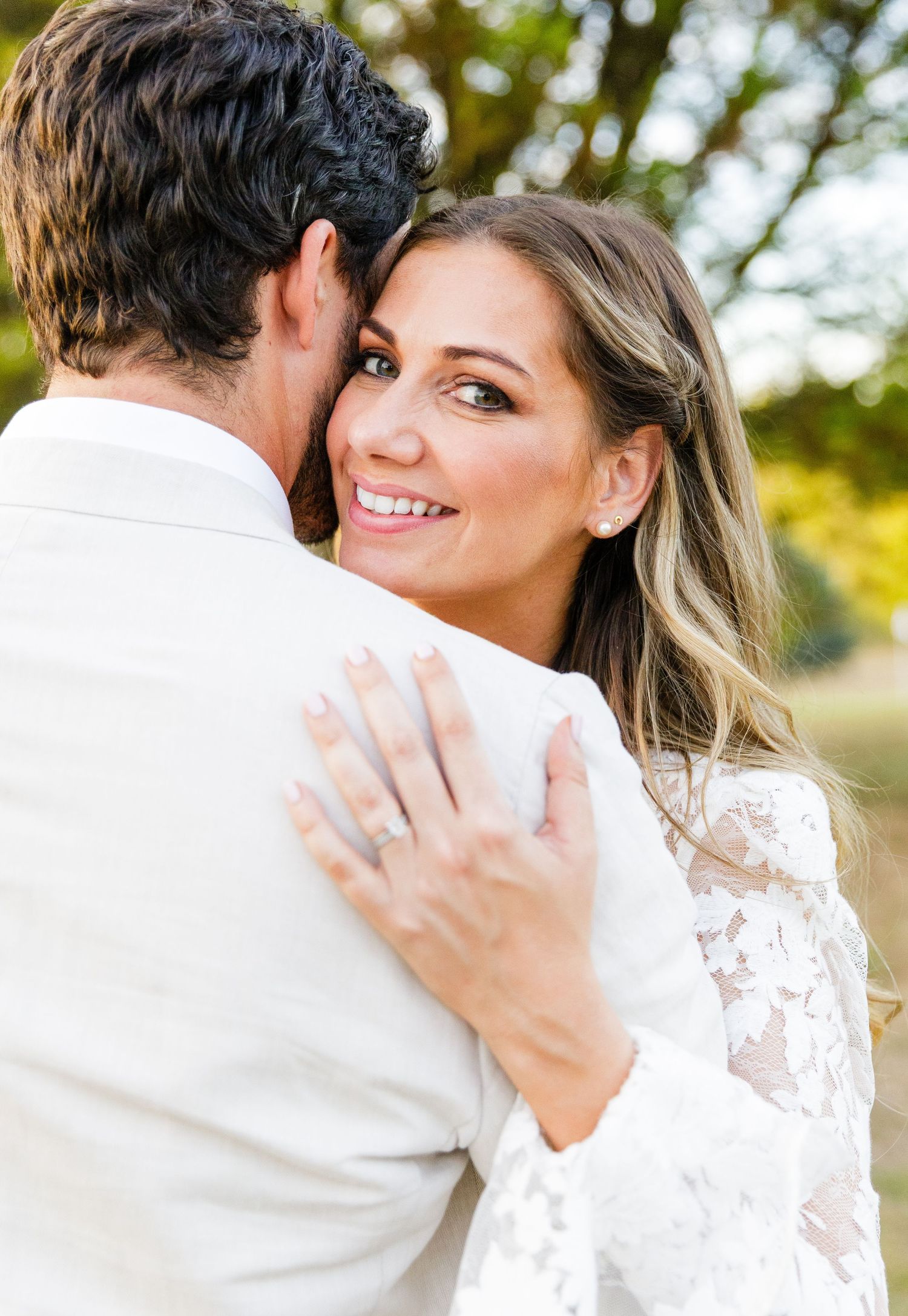 A romantic embrace between a couple in white, with sunlit trees creating a dreamy outdoor backdrop.