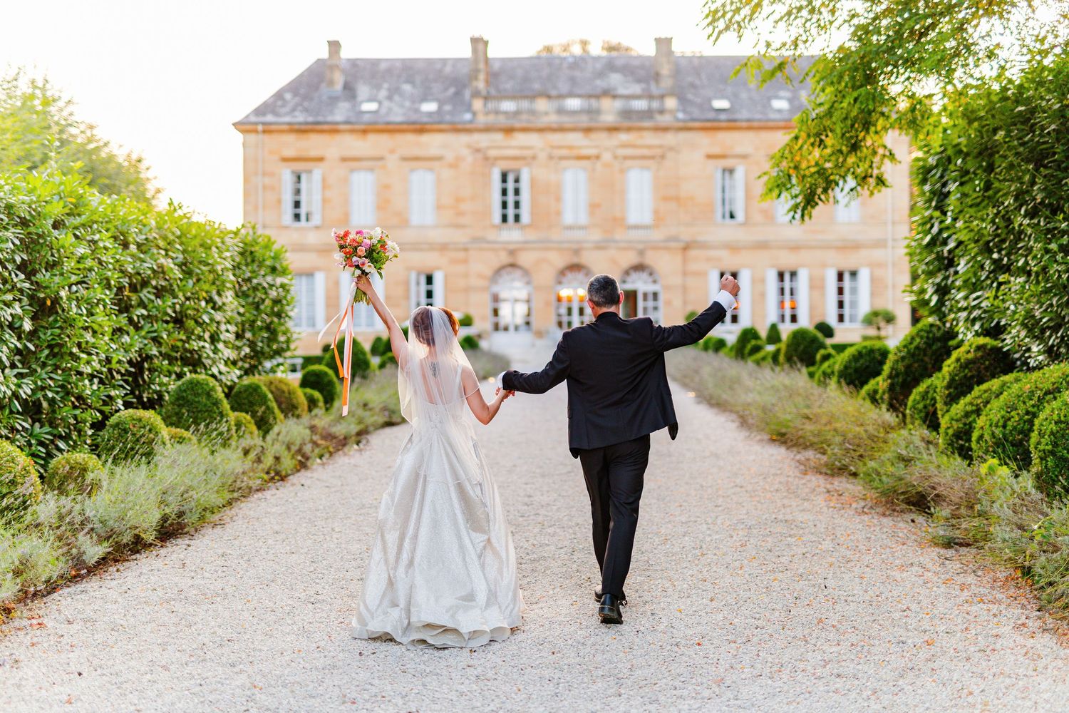 Bride and groom walking hand-in-hand toward a French château, raising their arms in celebration on a tree-lined gravel path.