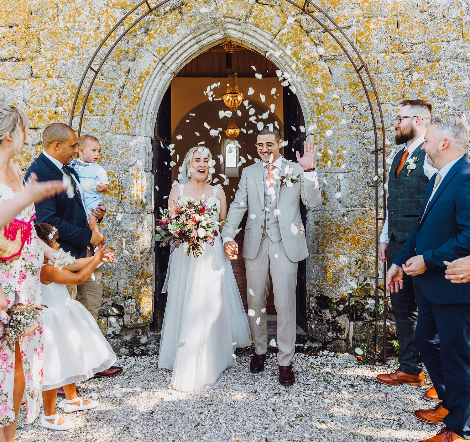 Wedding celebration with confetti throw at historic stone church archway as couple exits during joyful ceremony.